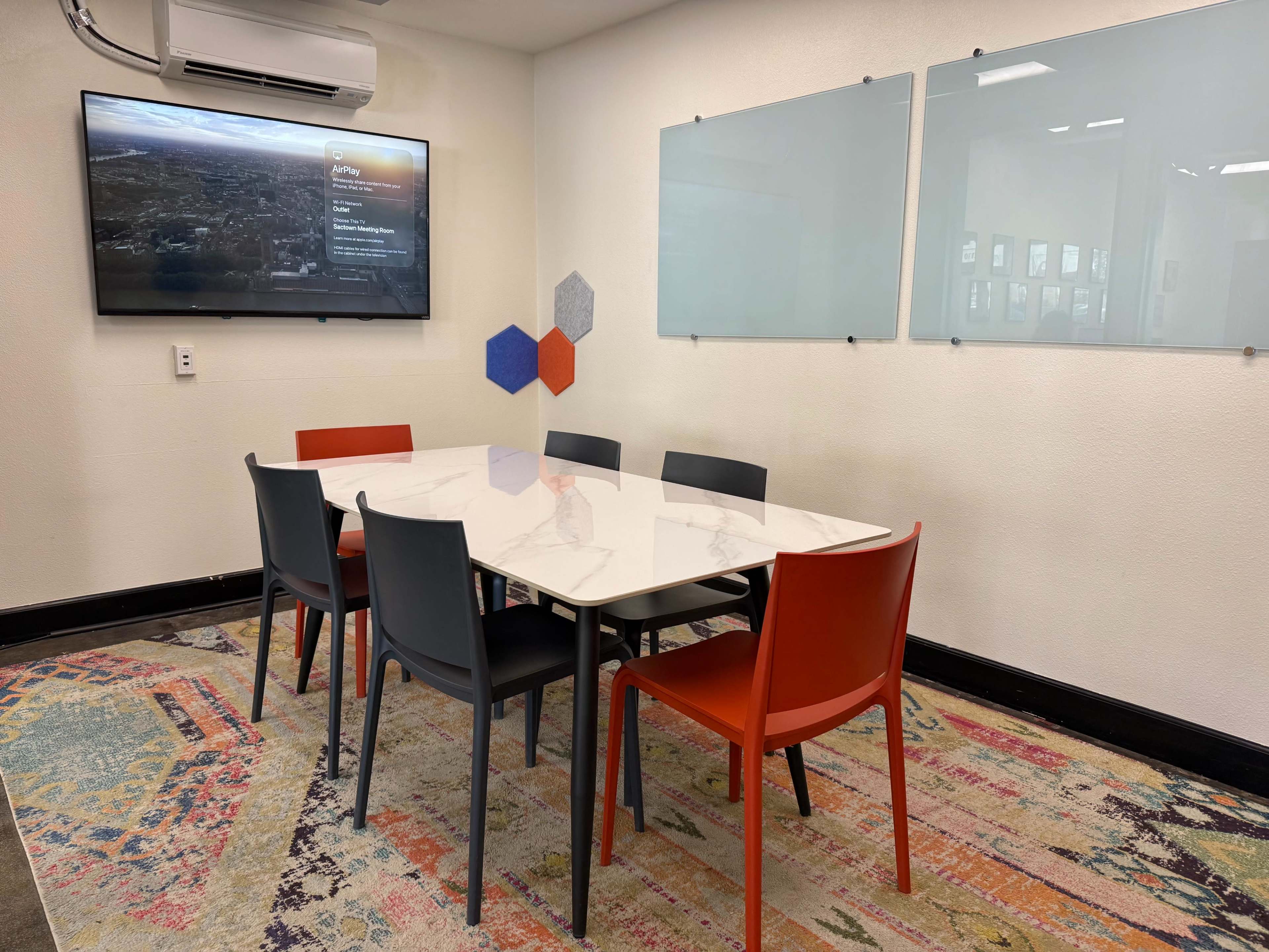 A conference room features a marble table surrounded by six chairs, with a large screen on the wall and a colorful rug underneath.