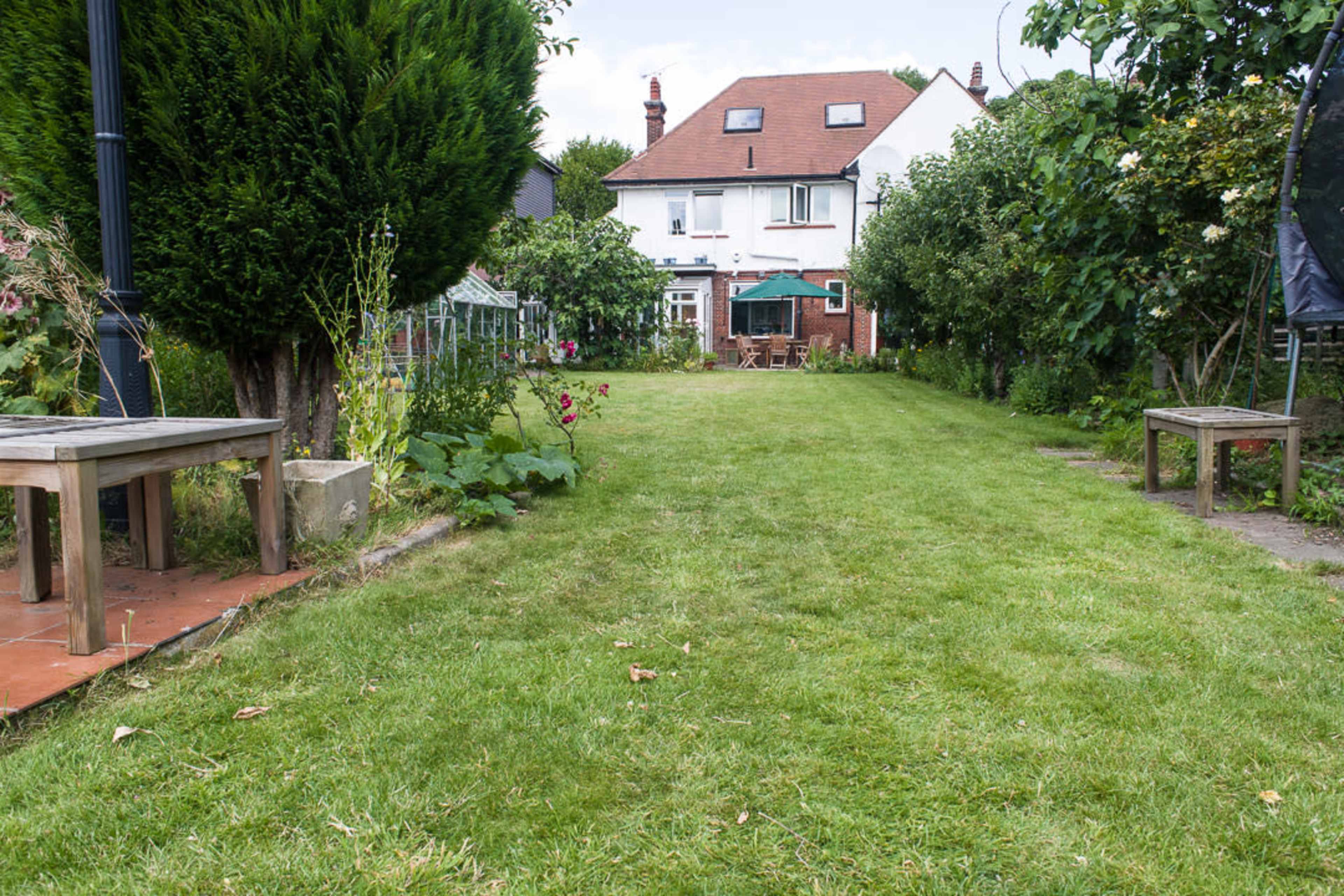 A well-maintained backyard features a long grassy lawn leading to a house with a red roof, surrounded by trees and garden structures.