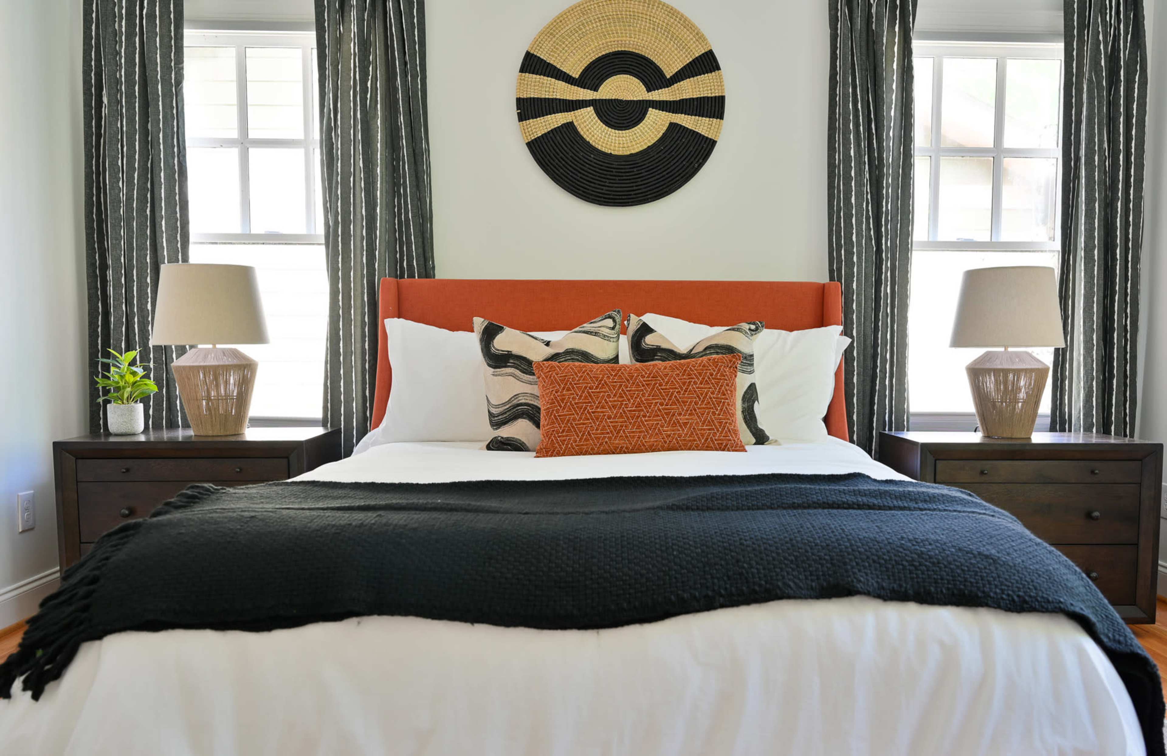 A neatly arranged bedroom featuring a bed with orange and black accents, flanked by wooden nightstands with lamps and decorated with a large circular wall hanging above the headboard.