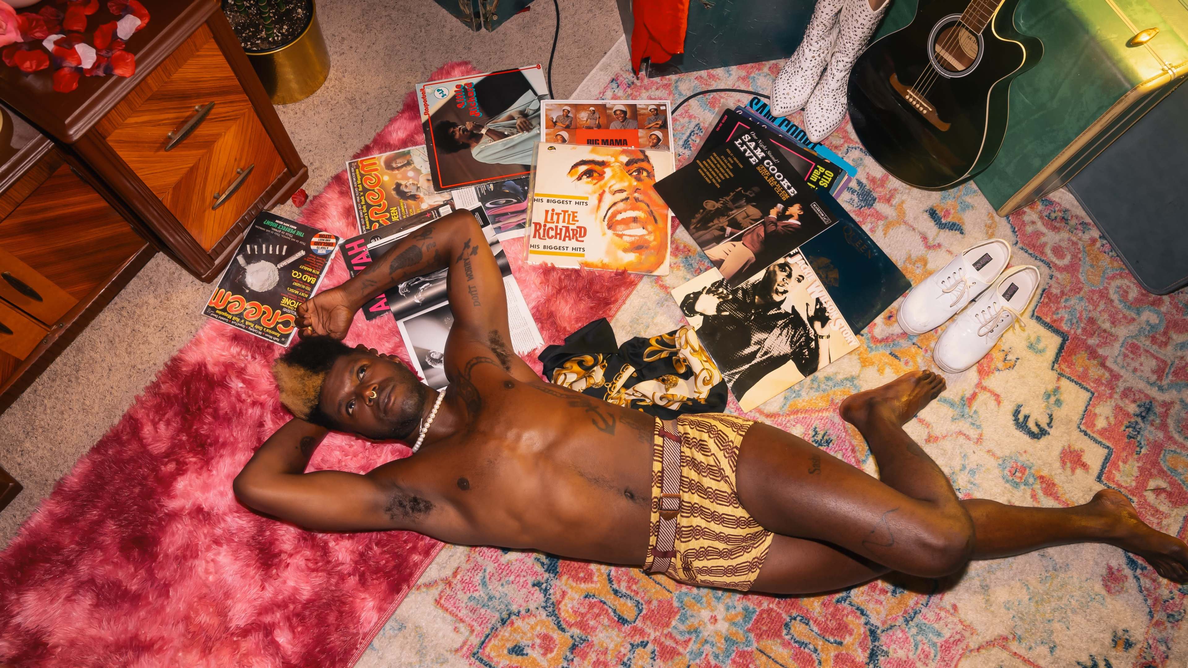 A person lying on a colorful rug surrounded by vinyl records and a guitar in a cozy room.