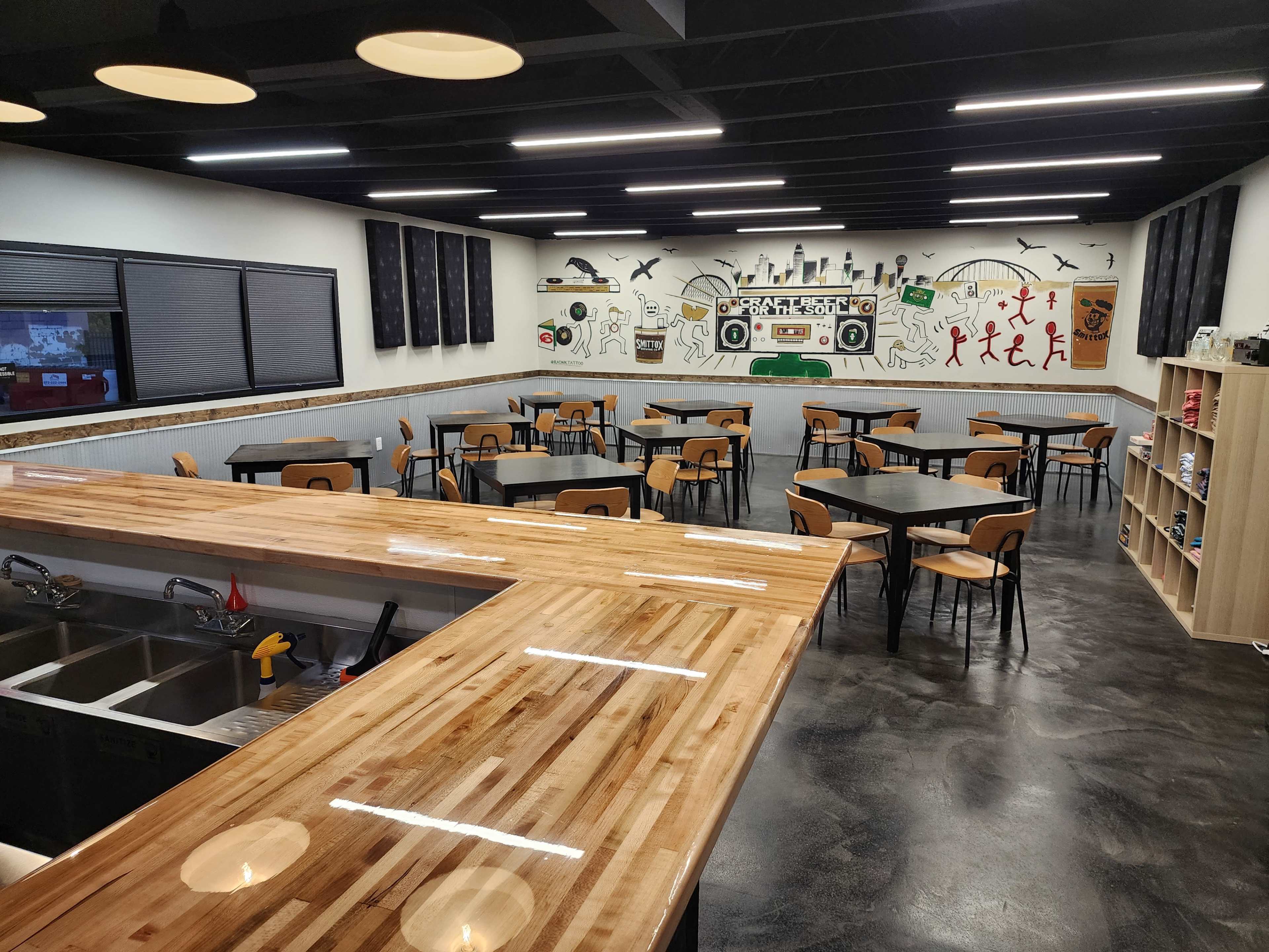 The image shows a modern, spacious cafe with several wooden tables and chairs arranged neatly, a long wooden counter, and a wall decorated with various illustrations.