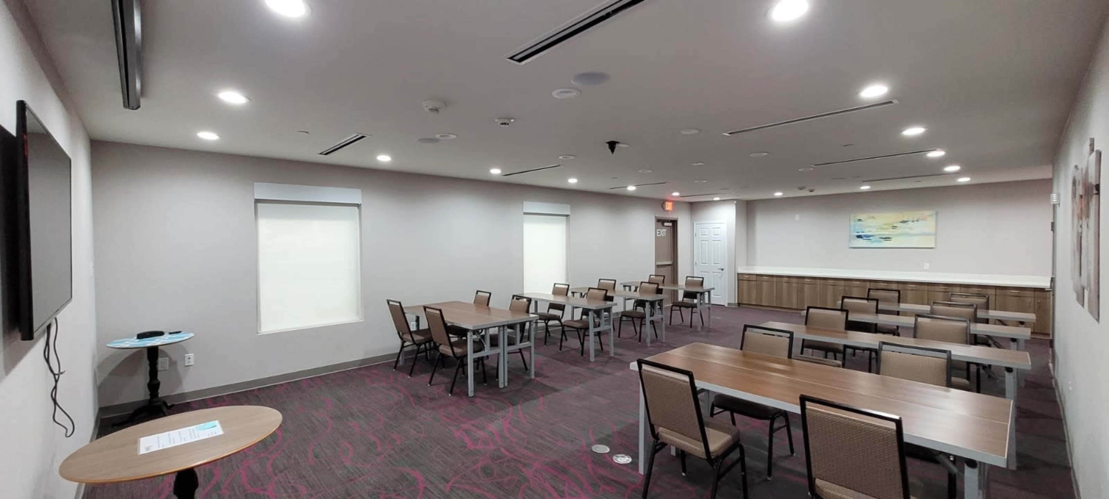 The image shows a spacious meeting room with multiple tables and chairs arranged neatly, along with a wall-mounted television and large windows.