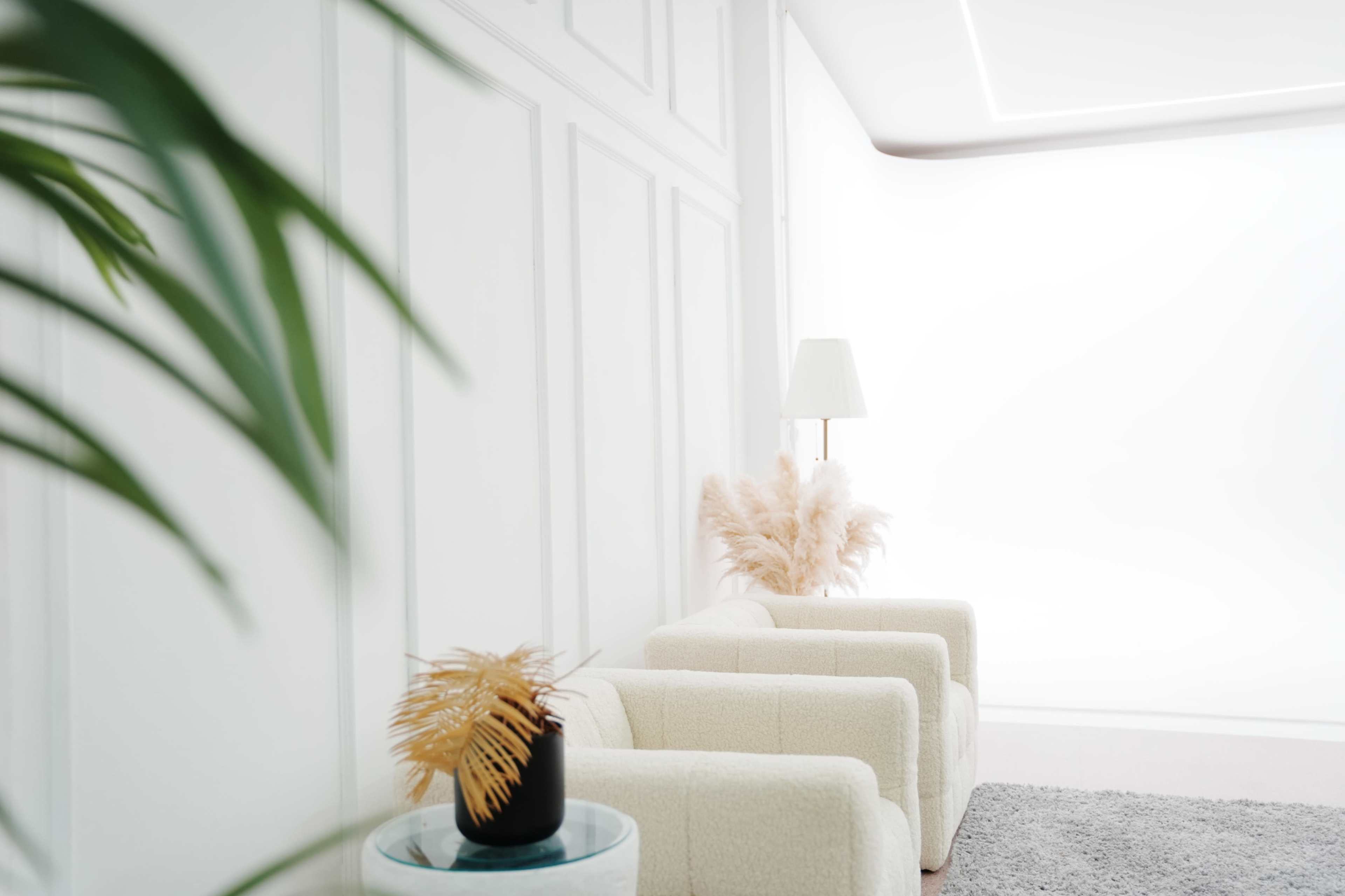 A minimalist living room features two white armchairs, a lamp, and a glass table with a plant and decorative vase against a light-colored wall.