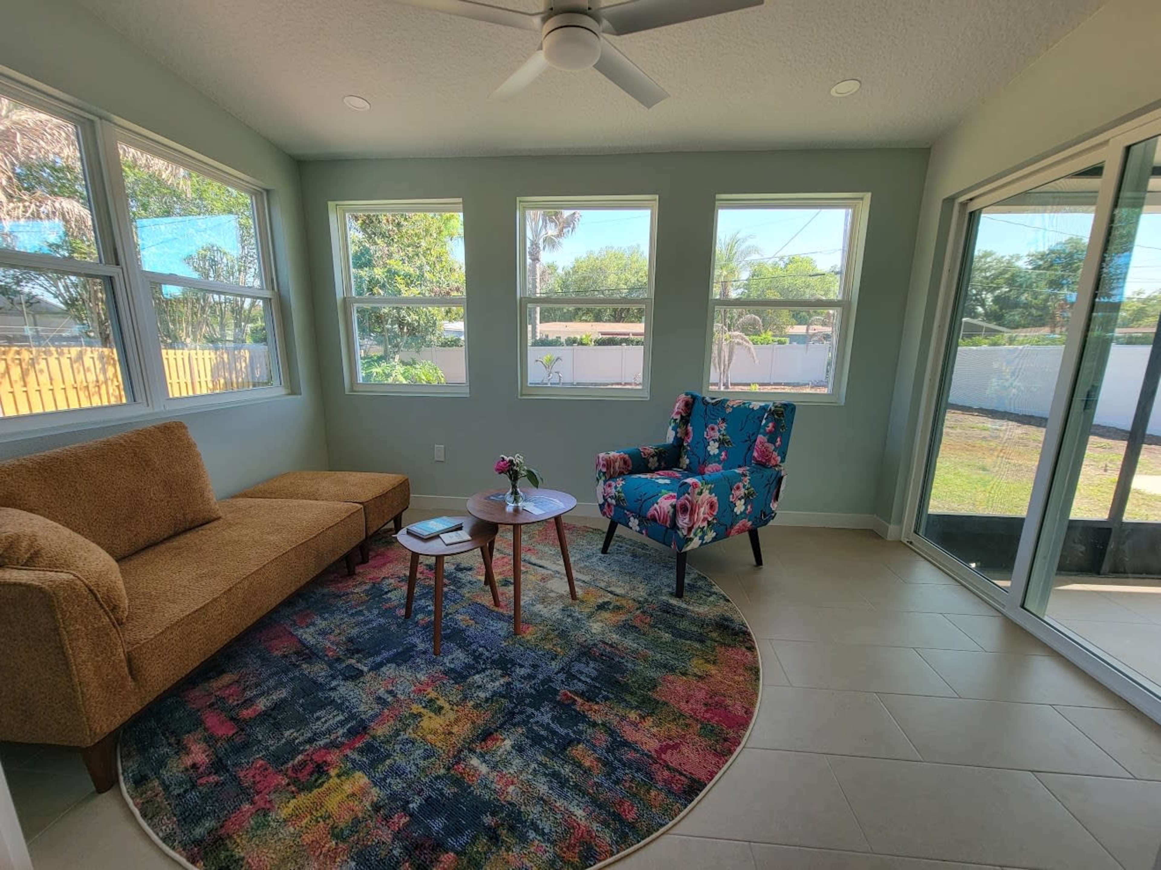 The image shows a bright sunroom featuring a brown sofa, a floral-patterned chair, a small table with a vase of flowers, and large windows that let in natural light.