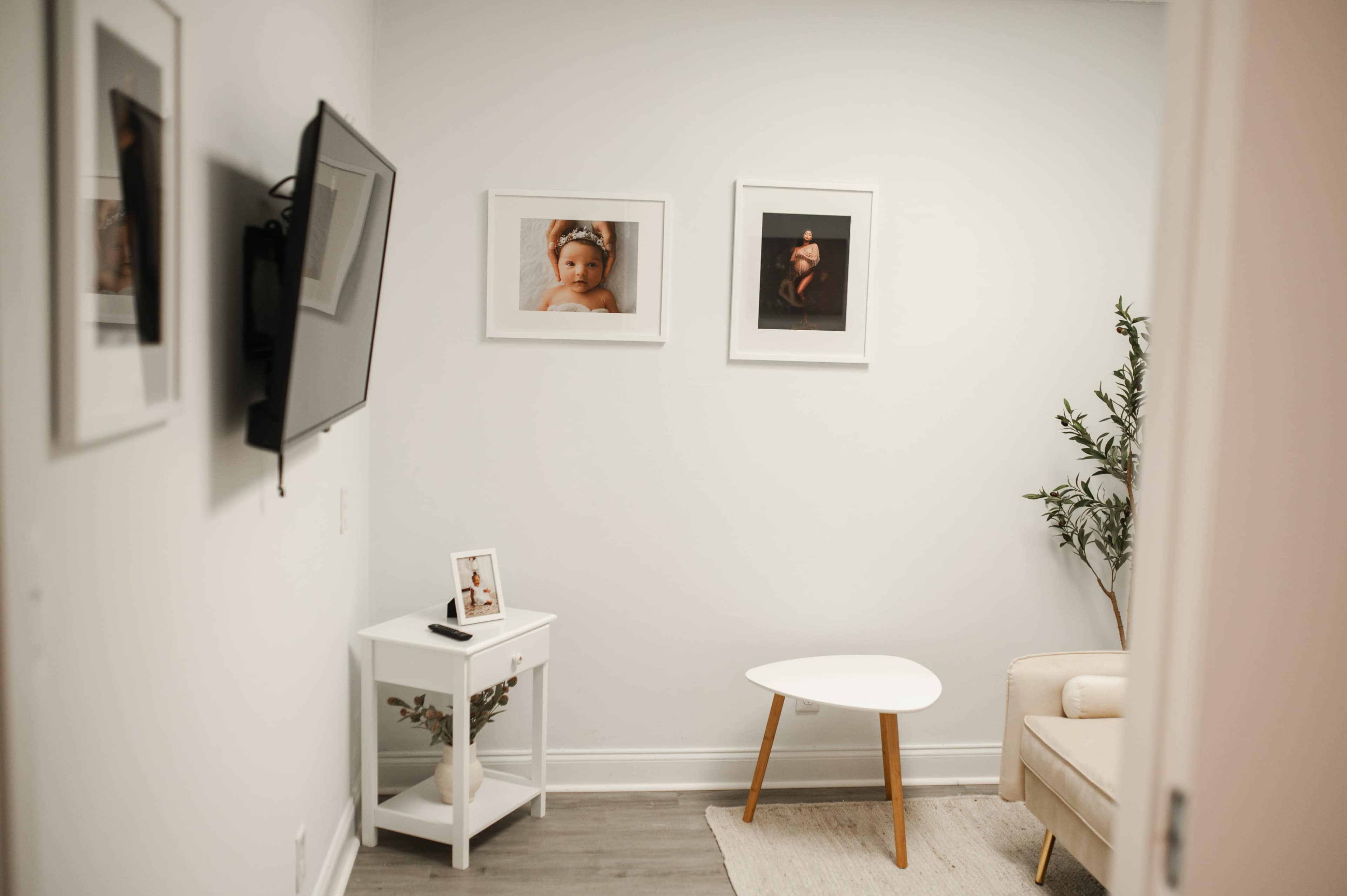 The image shows a small room with a TV mounted on the wall, framed photographs, a white side table, a plant, and a light-colored couch.