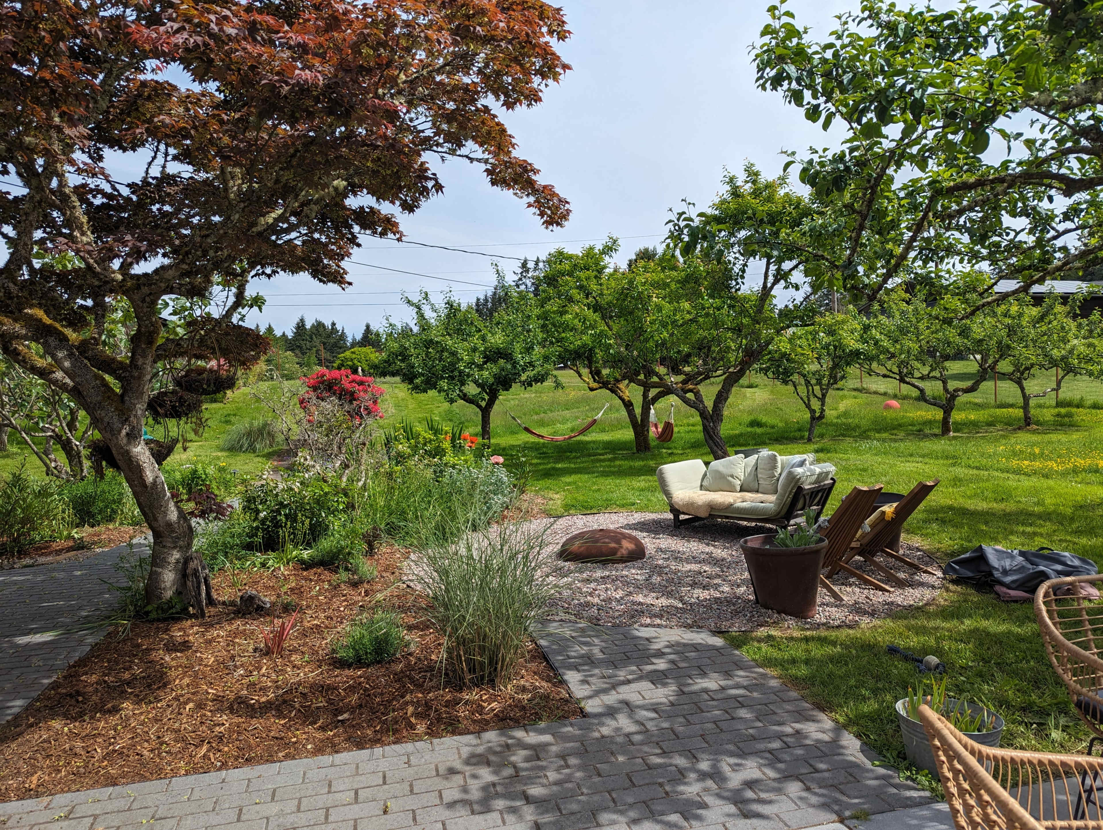 The image features a garden with a seating area, surrounded by trees and flowers, with a hammock visible in the background.