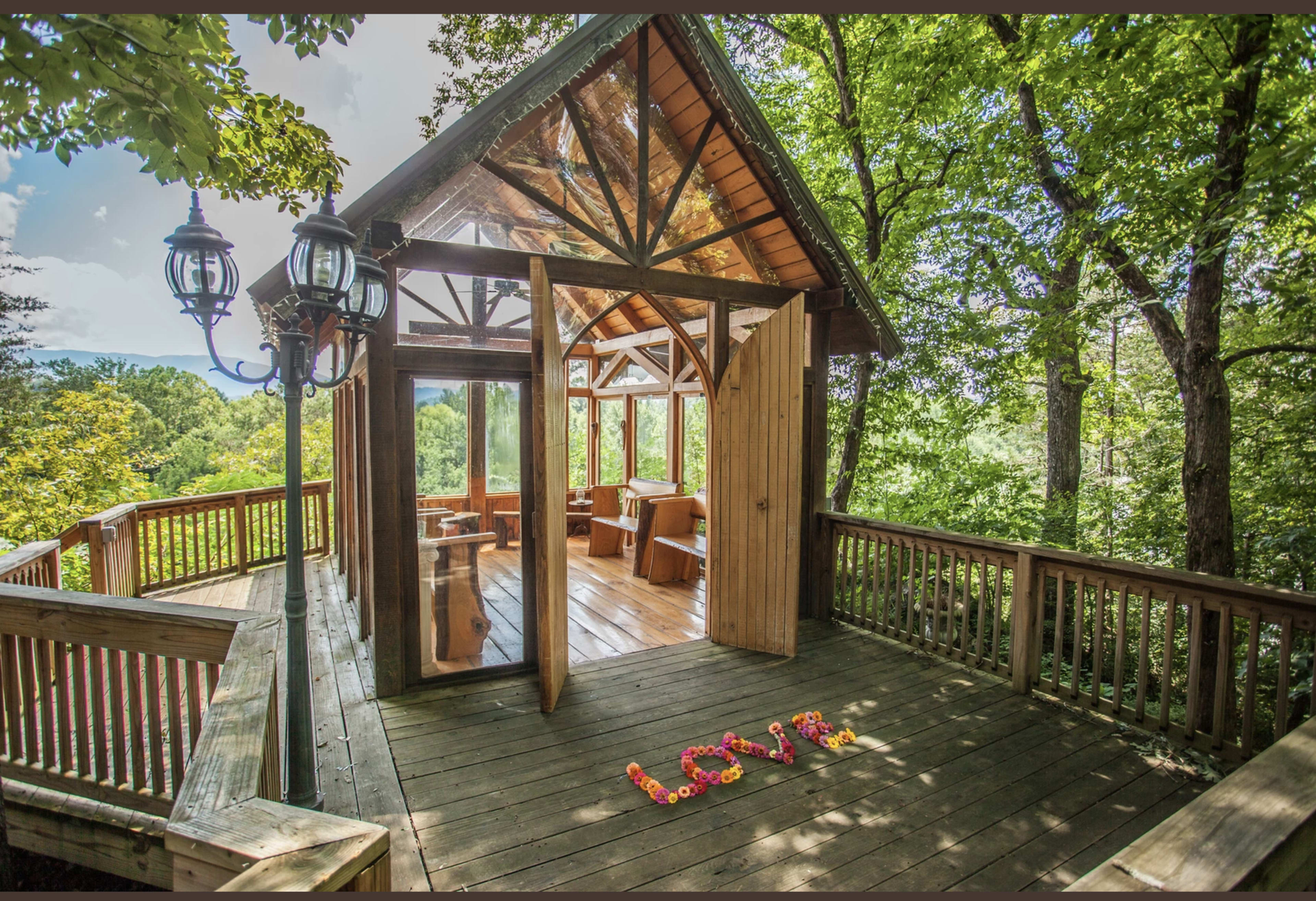 A wooden deck features a glass-paneled structure surrounded by trees, with the word "LOVE" spelled out in colored flowers on the floor.