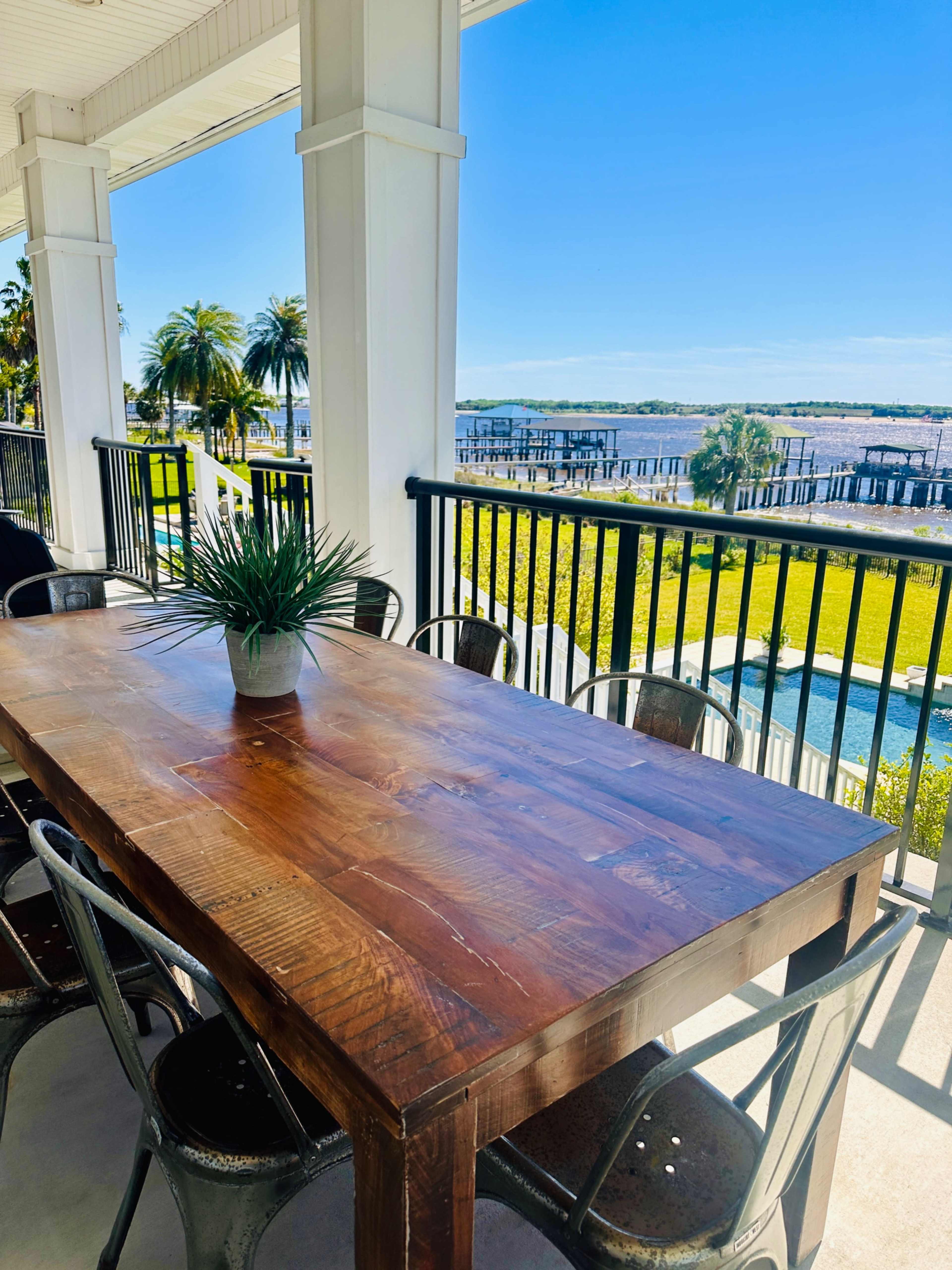 A wooden dining table with metal chairs is set on a porch overlooking a pool and landscaped area with palm trees and a waterfront view.