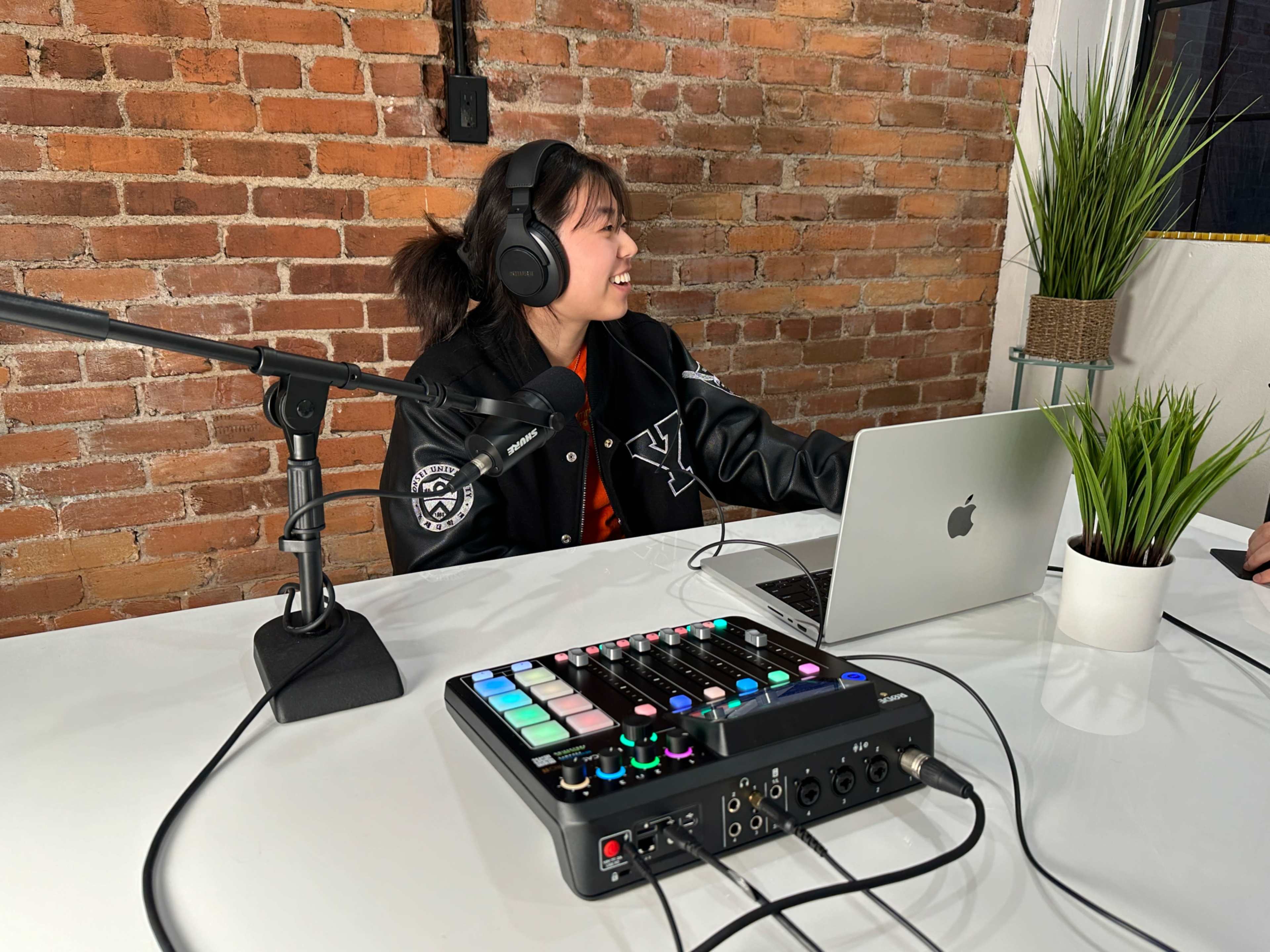 A young woman wearing headphones sits at a table with a laptop and a music production console in a room with exposed brick walls.