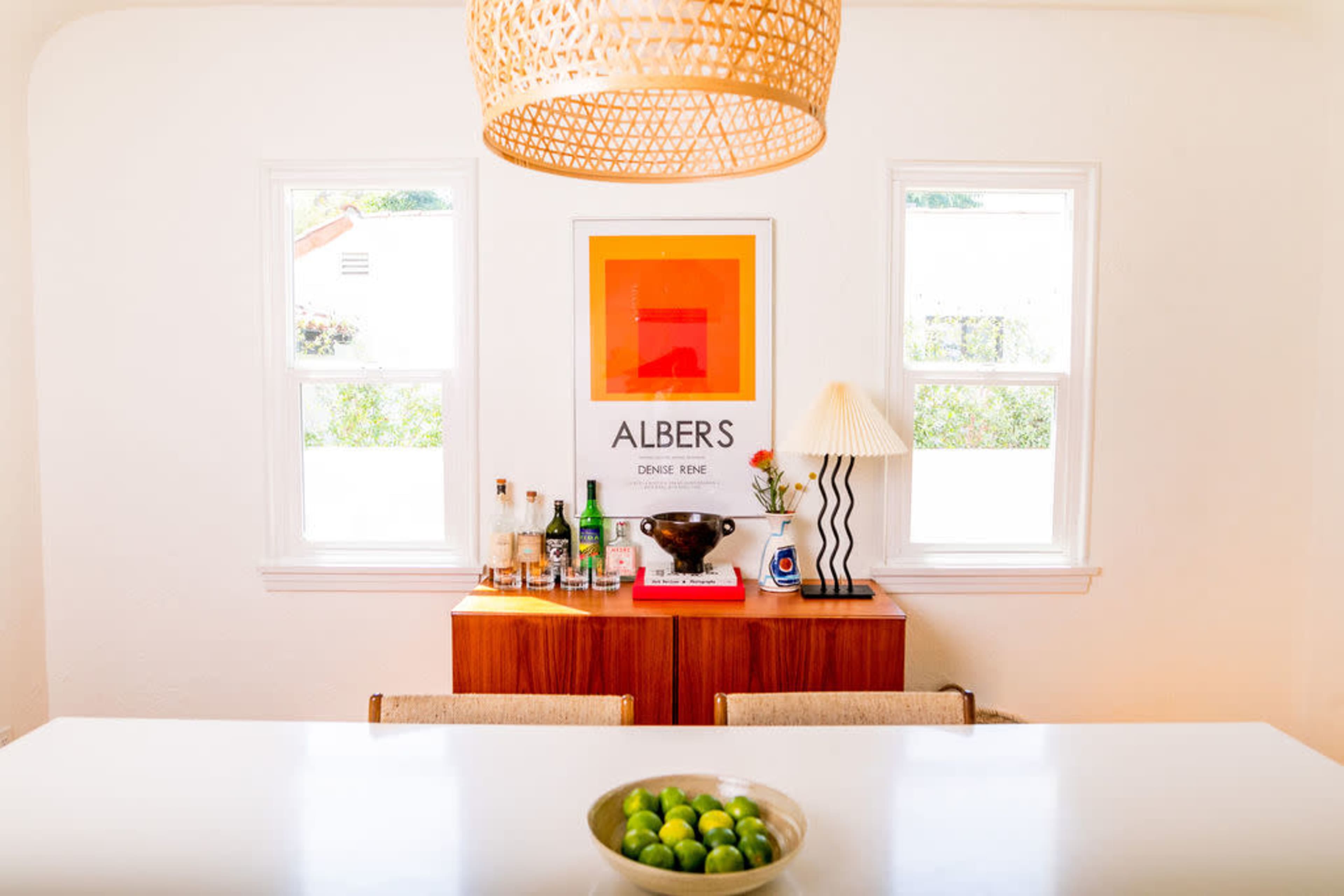 A modern dining area features a white table with a bowl of green limes, a wooden sideboard displaying various bottles and a black bowl, and a large framed artwork on the wall.