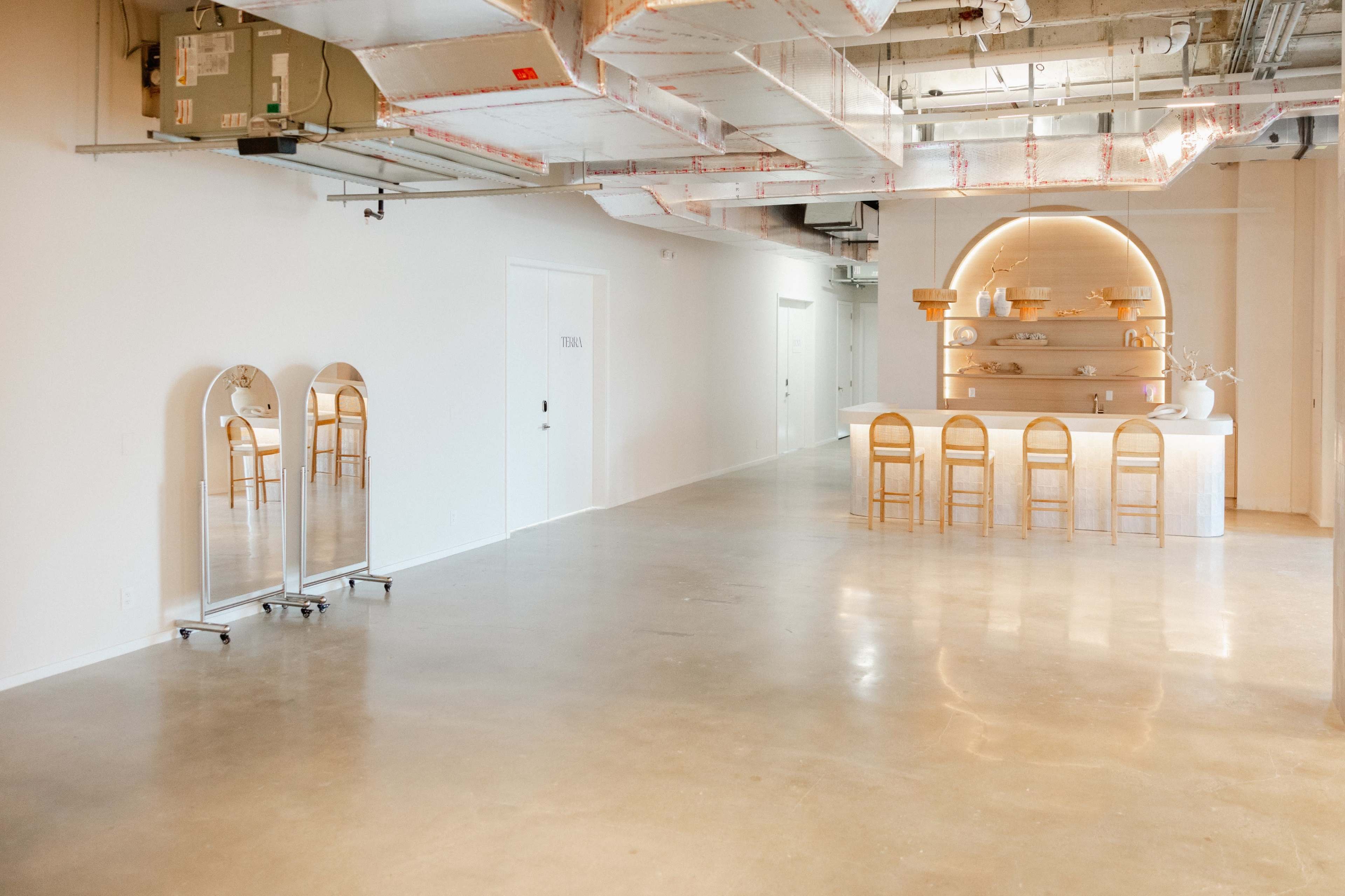 The interior of a modern, minimally designed space features a bar with stools at the far end, illuminated by soft, curved lighting, and mirrors along one wall.
