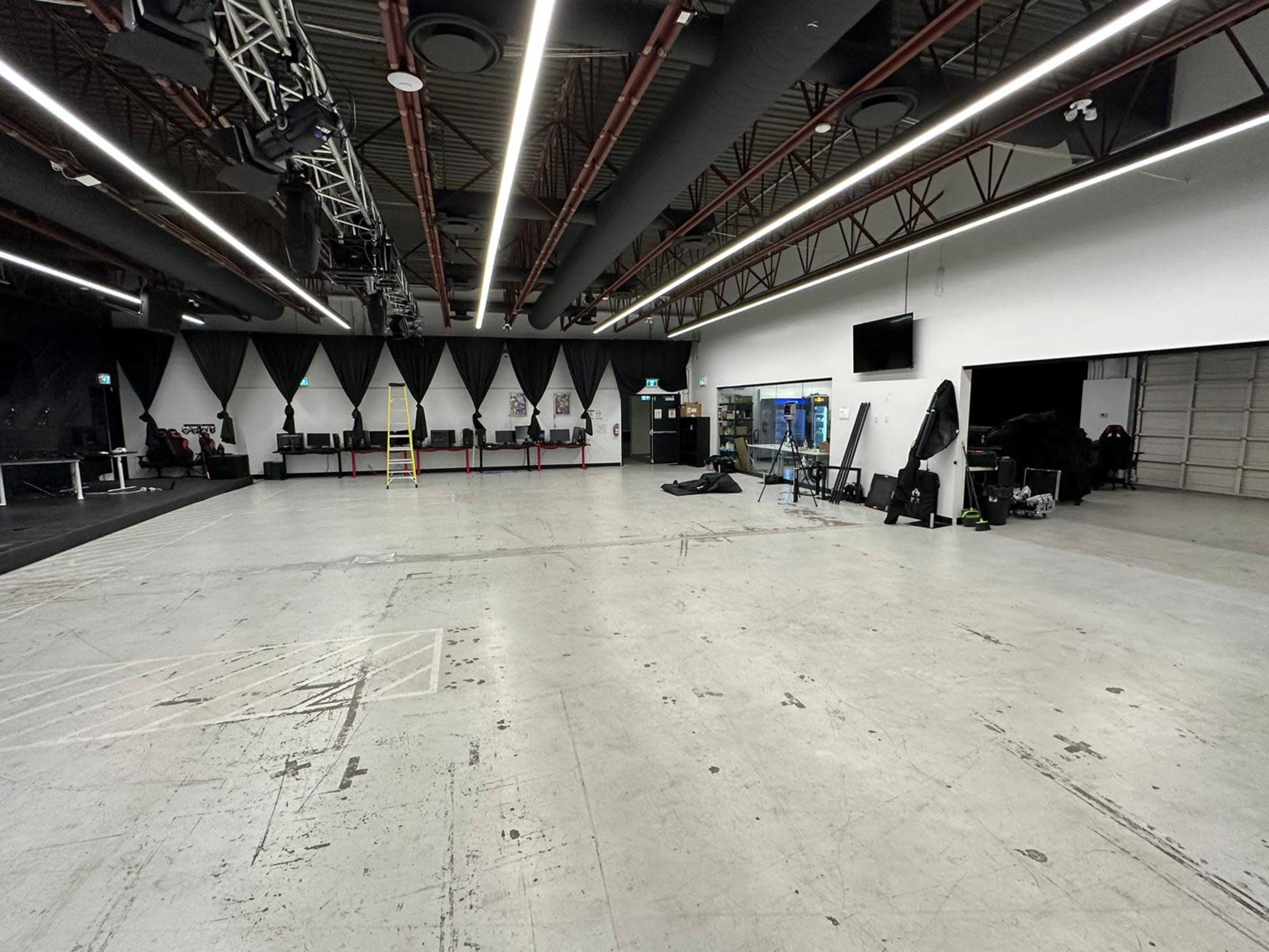 A spacious interior of a multipurpose studio with a concrete floor, white walls, and a mix of lighting and equipment visible.
