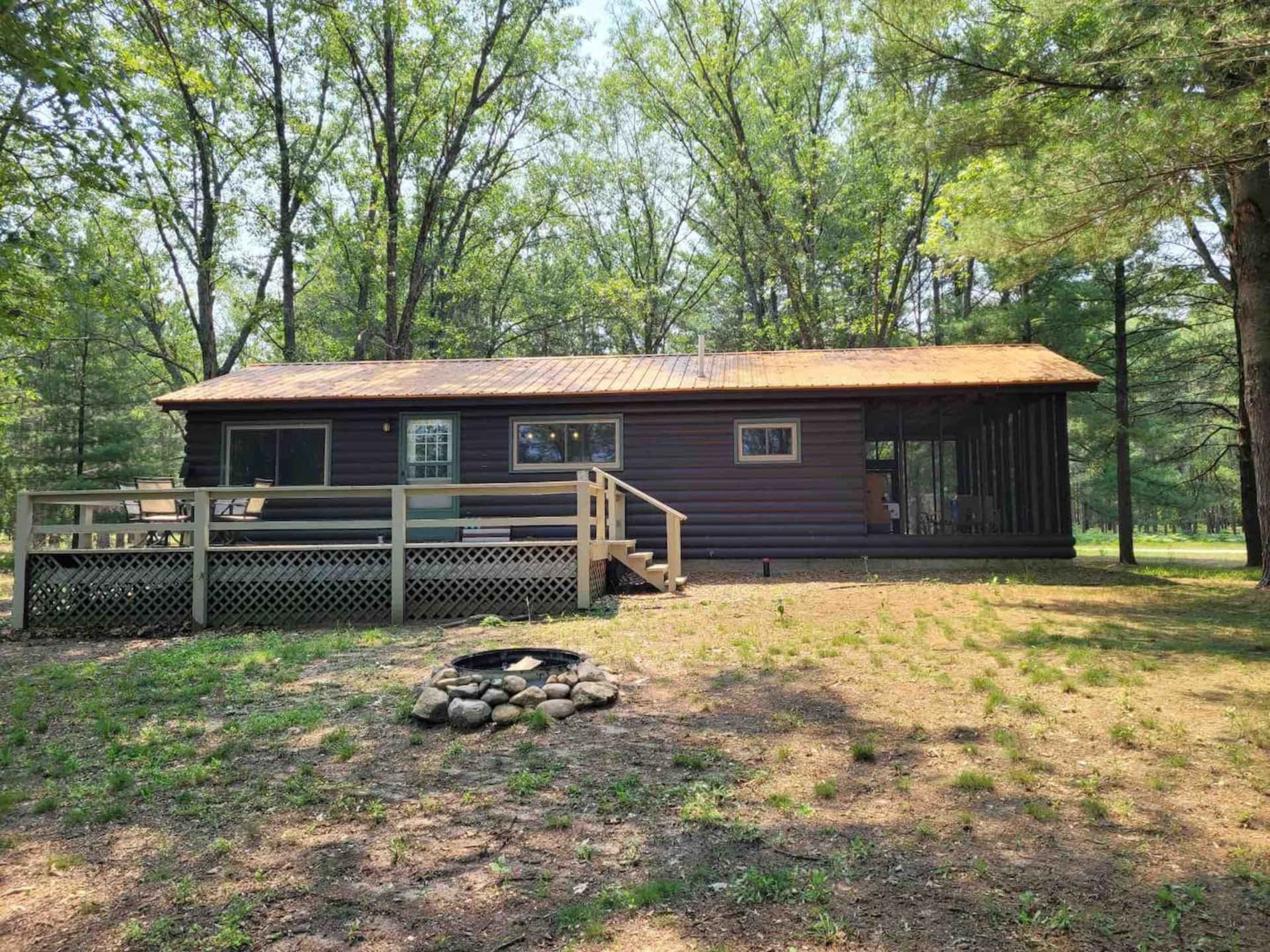 A rustic cabin with a wooden deck and a stone fire pit is surrounded by trees in a wooded area.