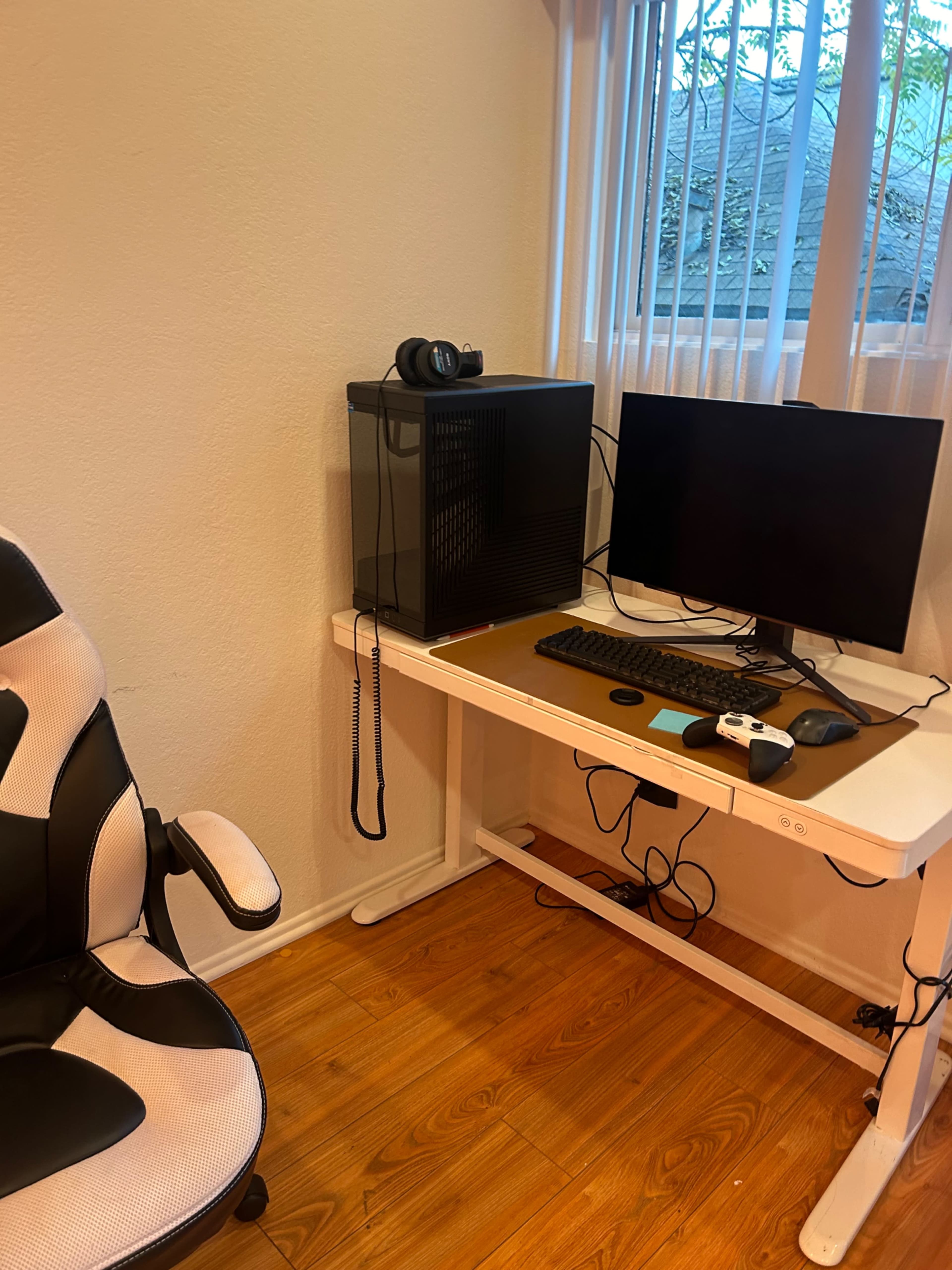 The image shows a sleek computer setup with a monitor, a gaming chair, and a black desktop PC on a wooden desk against a wall with window blinds.