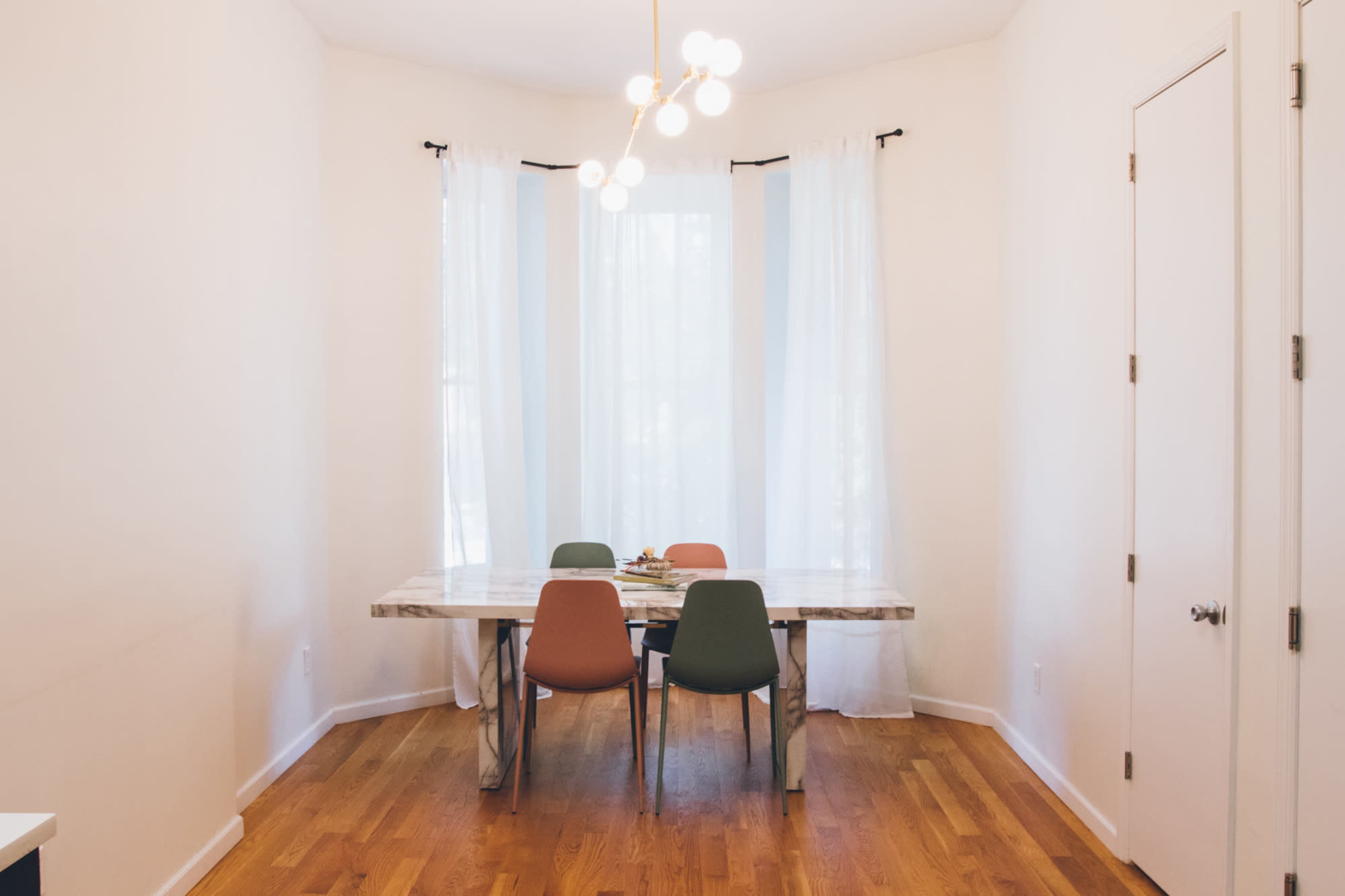 A dining area features a marble-top table surrounded by four chairs in a room with large windows and sheer white curtains.