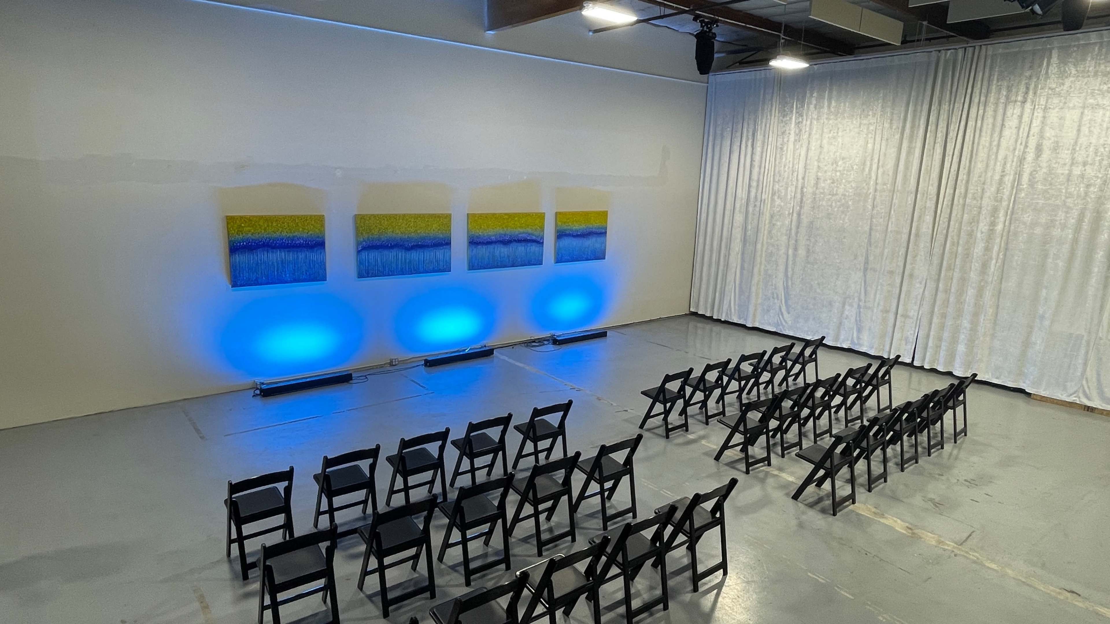A spacious room features black chairs arranged in rows facing three large paintings illuminated by blue lights against a curtain backdrop.