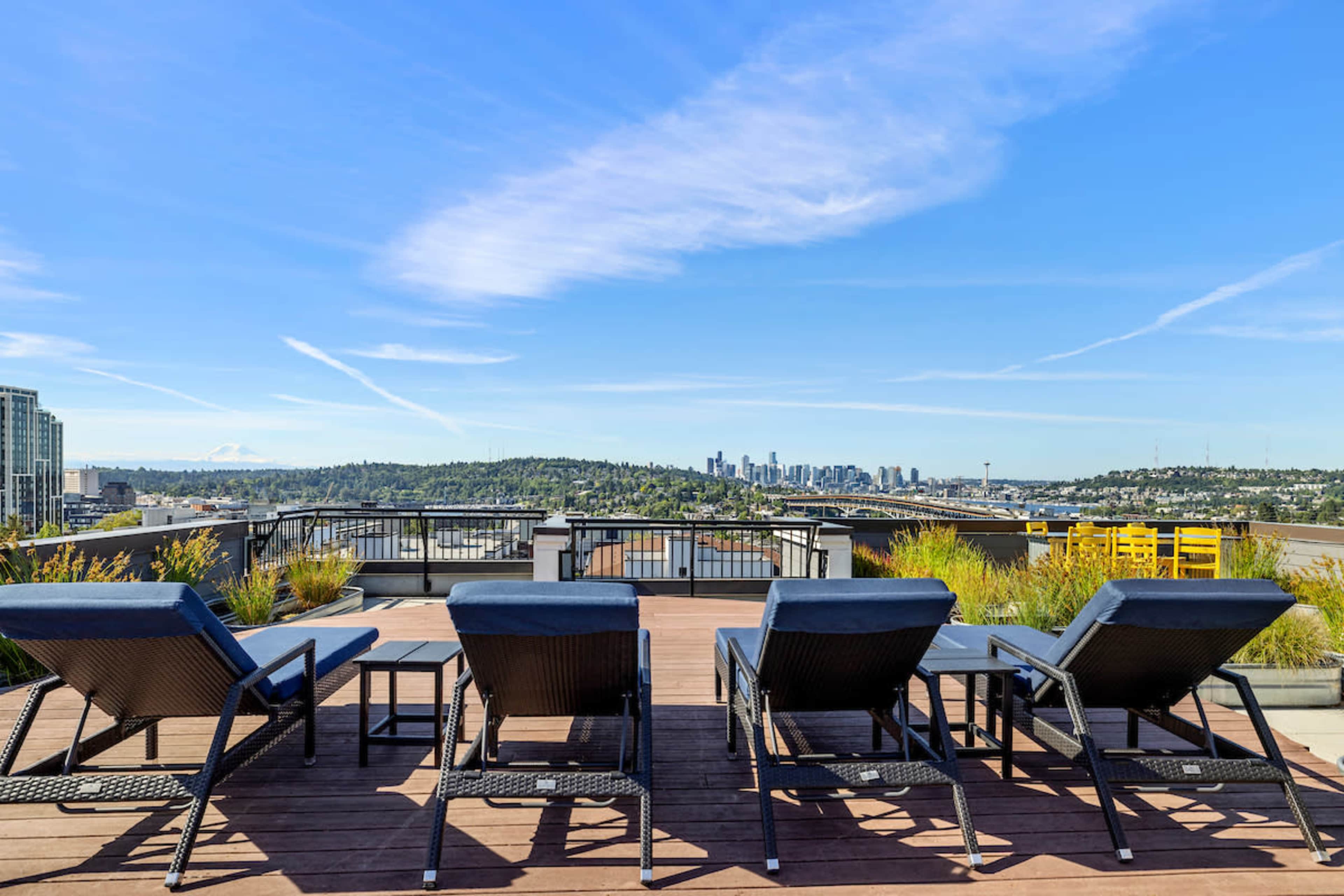 The image shows a rooftop terrace with several lounge chairs facing a panoramic view of a city skyline against a clear blue sky.