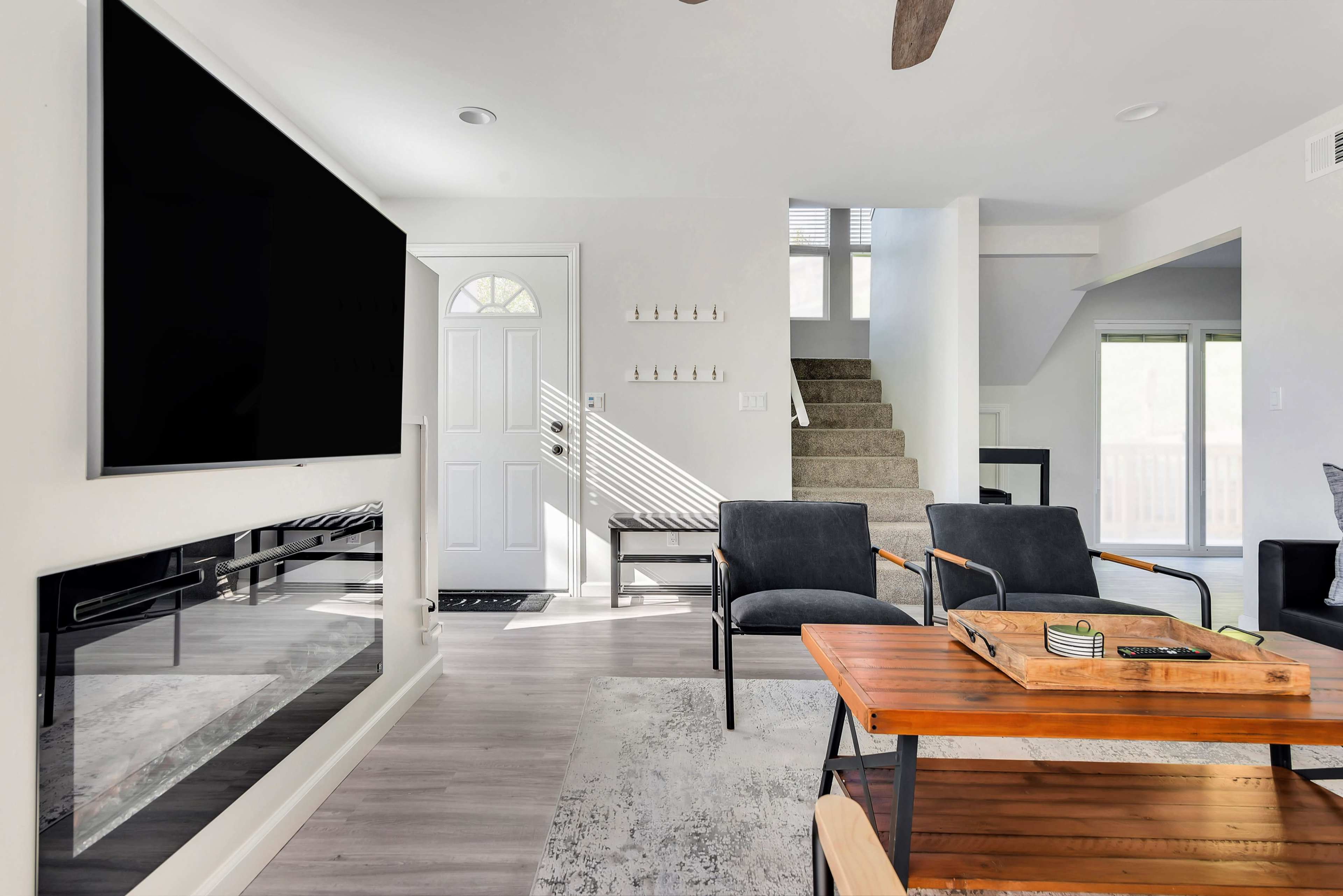 A bright living room featuring a modern television mounted on the wall, a wooden coffee table with a tray, and a staircase in the background leading to an upper level.