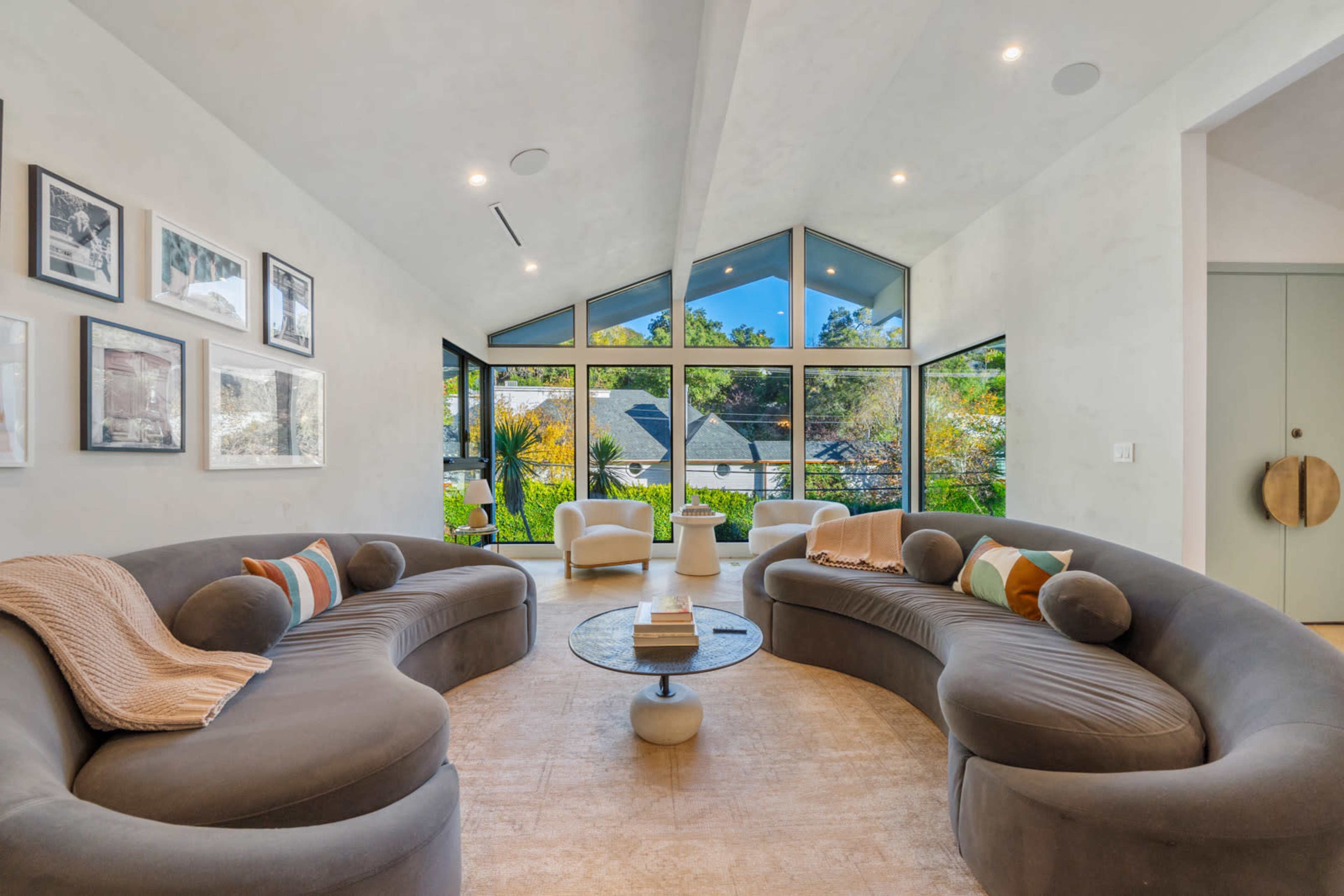 The image depicts a modern living room featuring two curved sofas facing a central coffee table, with large windows allowing natural light and a view of the outdoors.