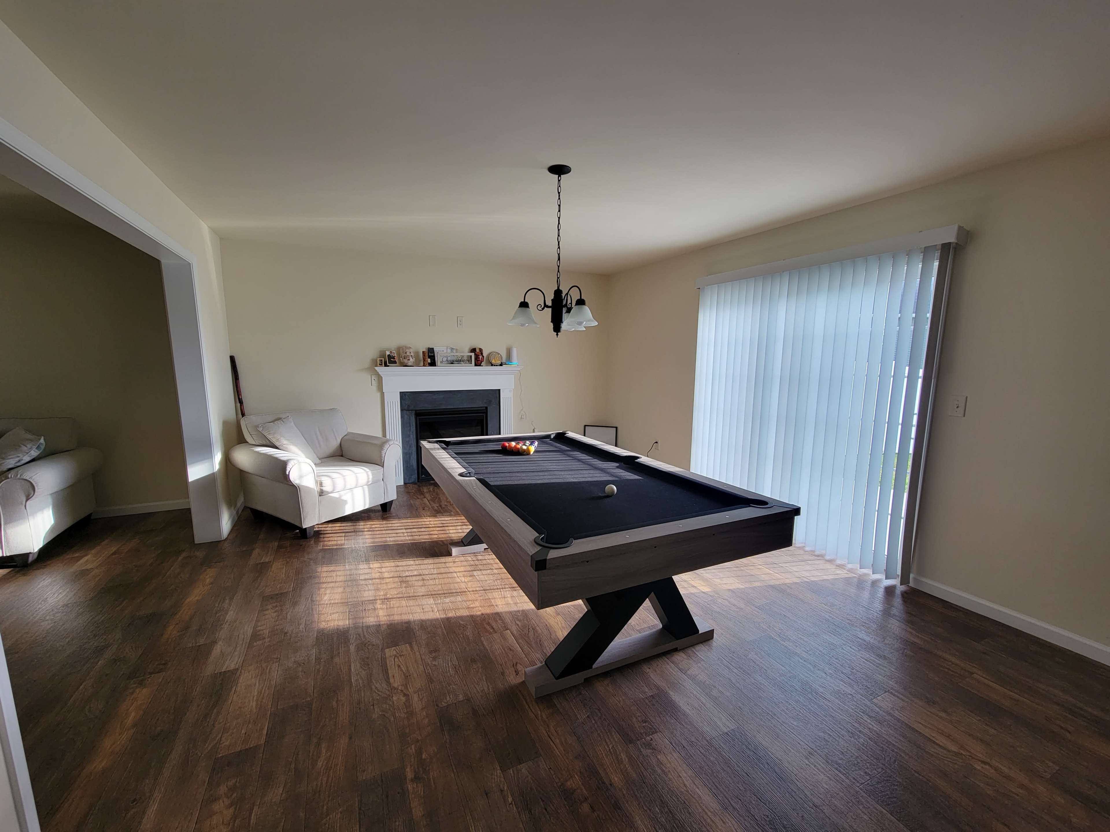 A spacious room featuring a billiard table, a fireplace, and two armchairs near a window with vertical blinds.