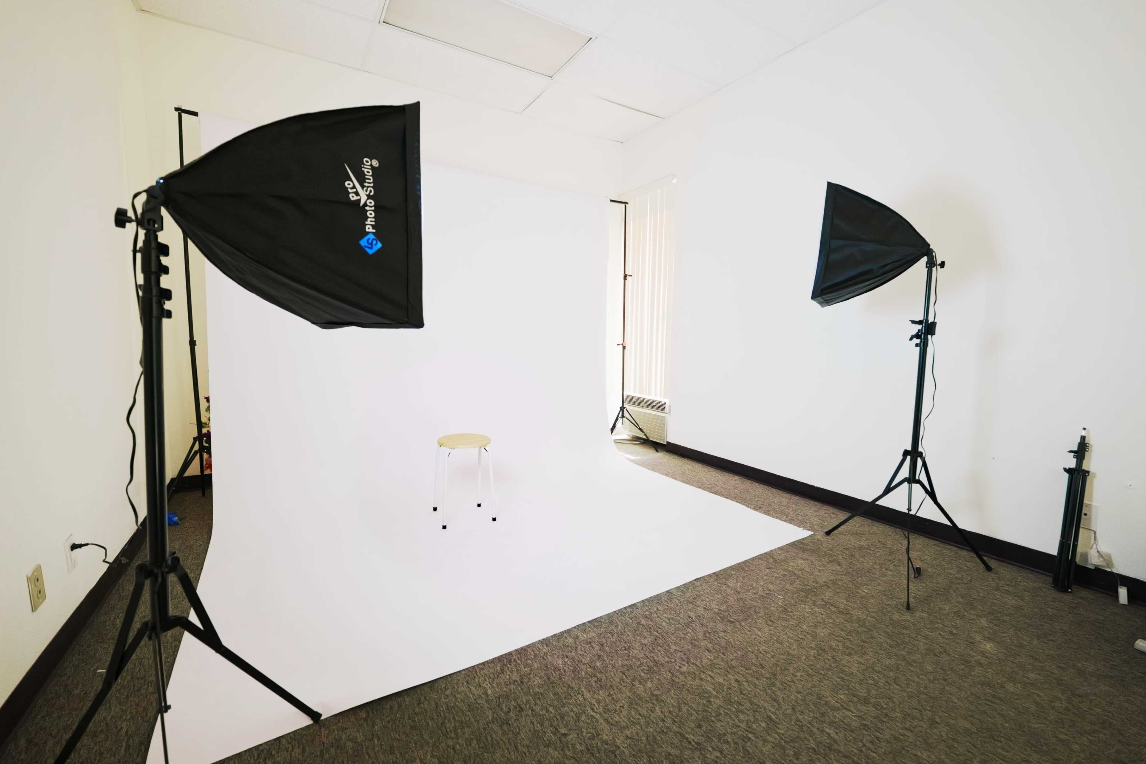 A photography studio is set up with two softbox lights facing a white backdrop and a single stool in the center.