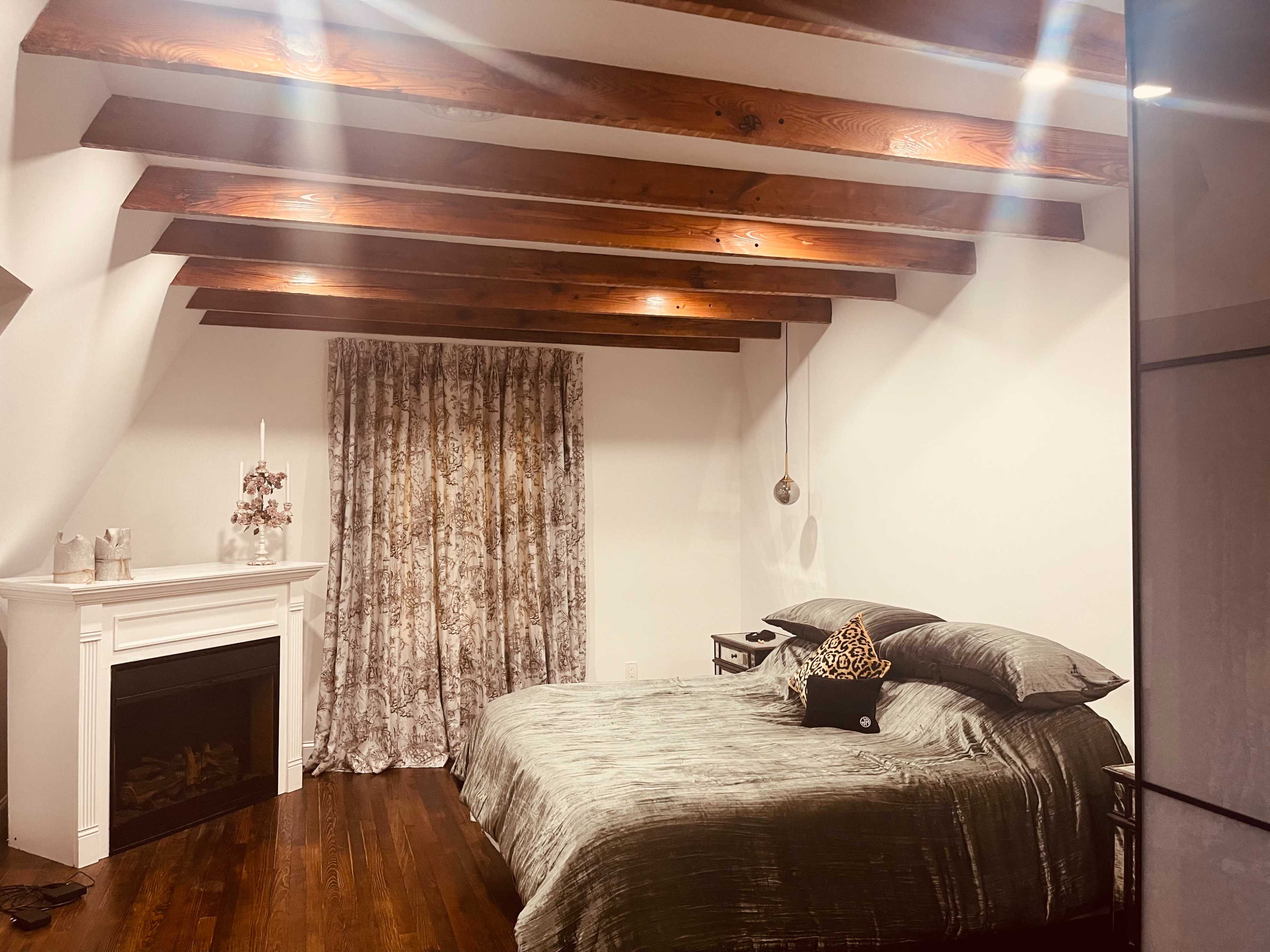 A bedroom features a large bed with gray bedding, rustic wooden beams overhead, and a window dressed with patterned curtains beside a decorative fireplace.