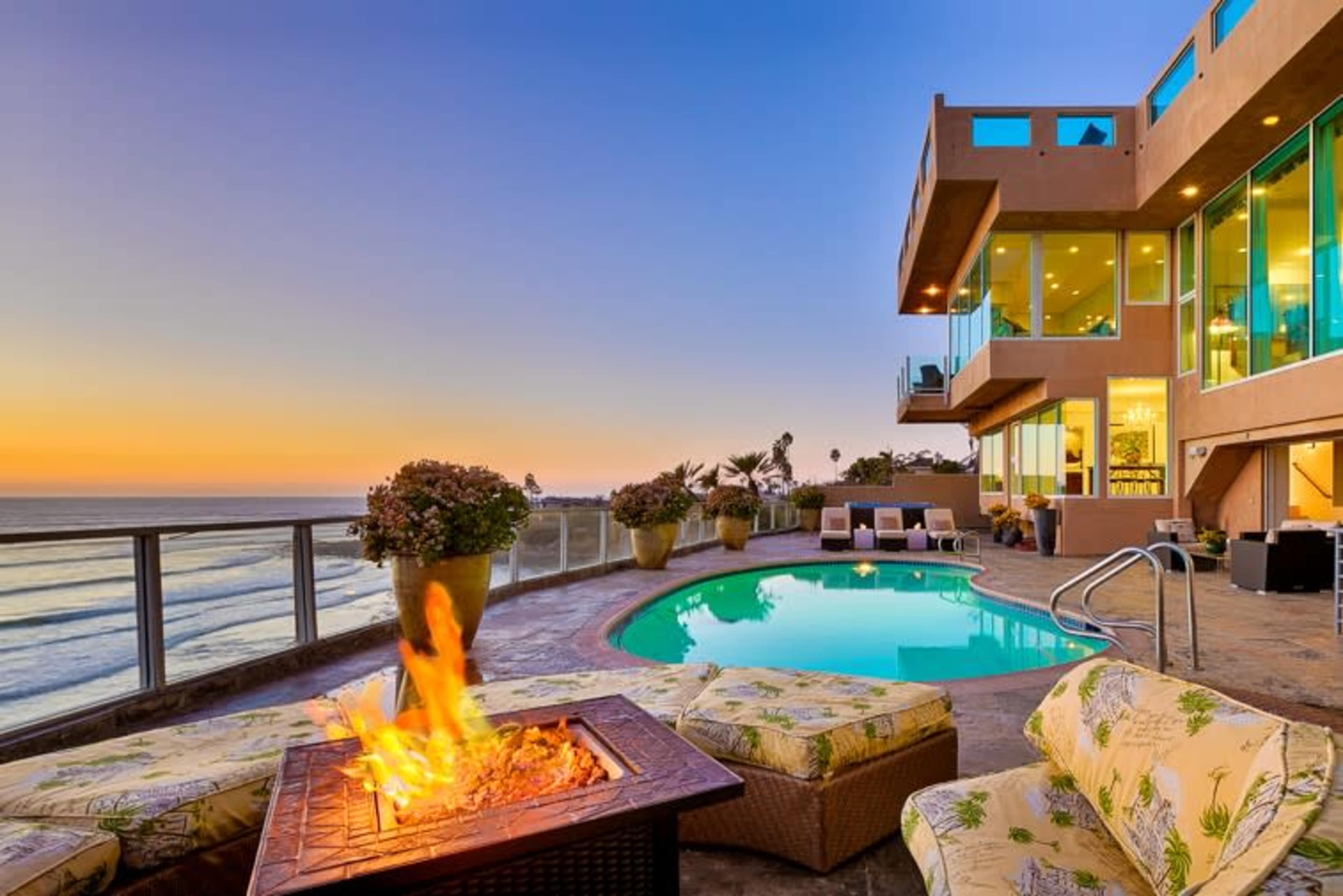 A modern home overlooks a pool with a fire pit, surrounded by seating, against a backdrop of a sunset over the ocean.
