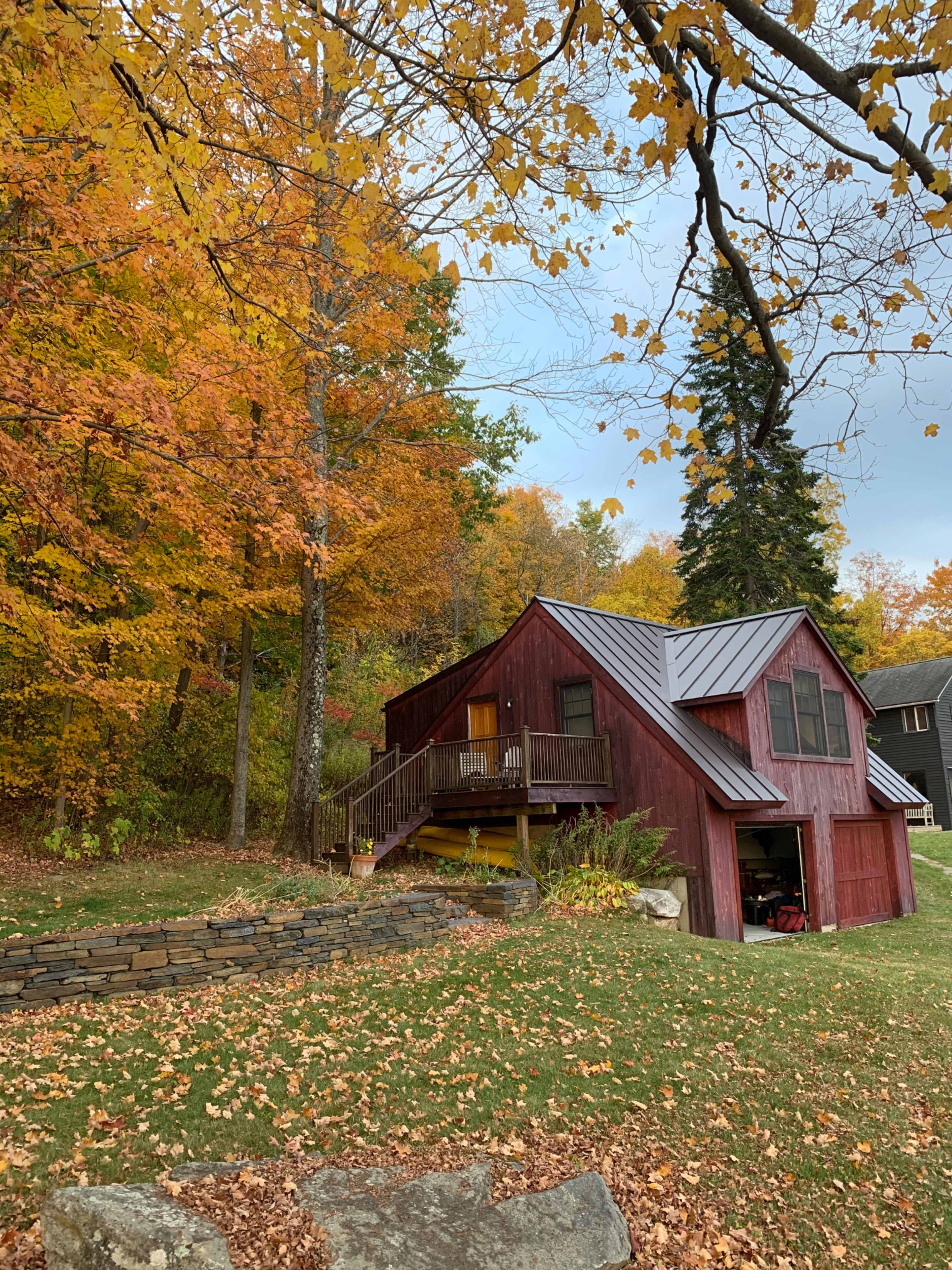 A red house with a metal roof is situated beside a grassy area surrounded by colorful autumn trees.
