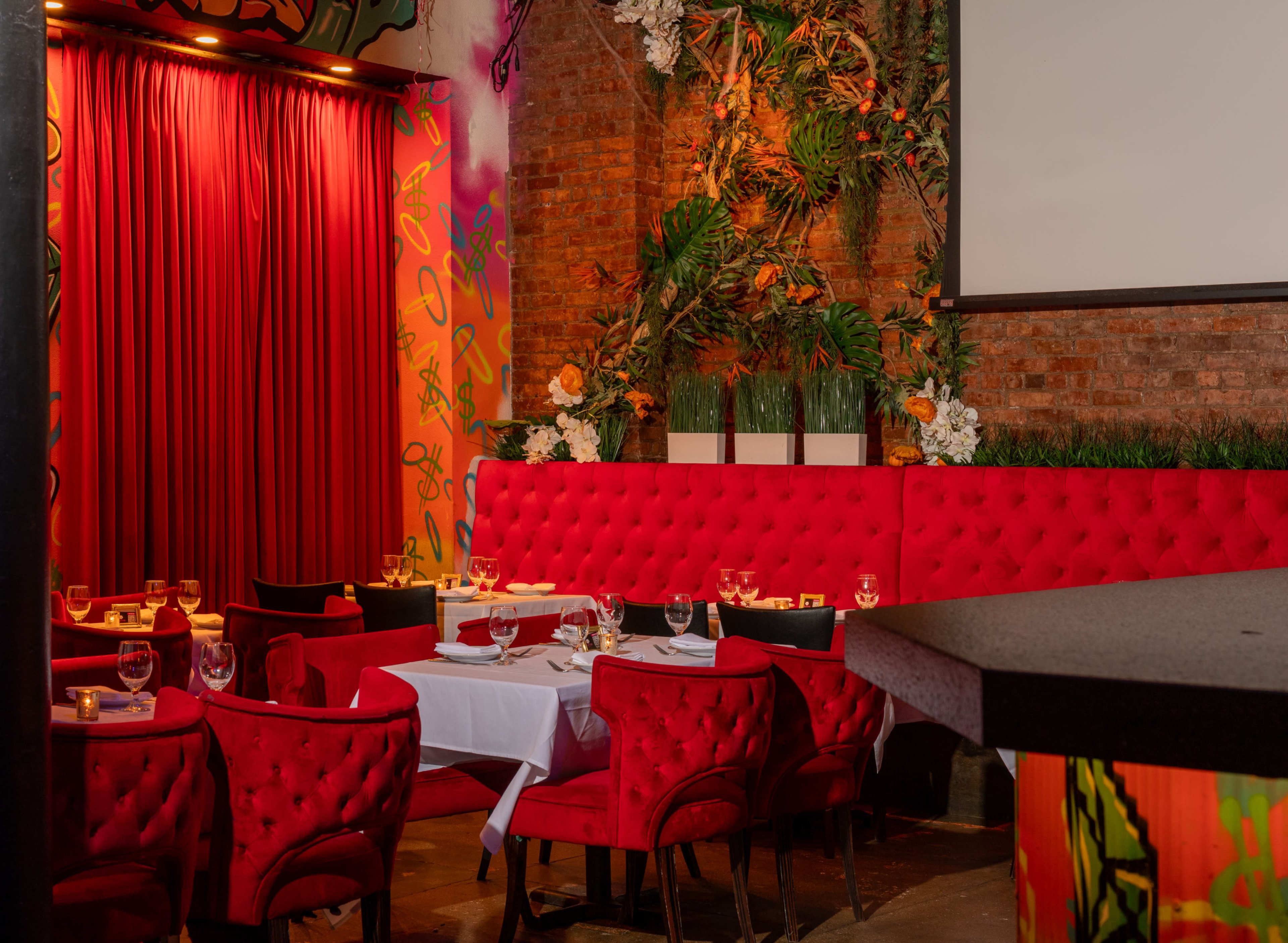 The image shows a vibrant restaurant interior with red velvet seating, white tablecloths, and a backdrop of colorful artwork and greenery.