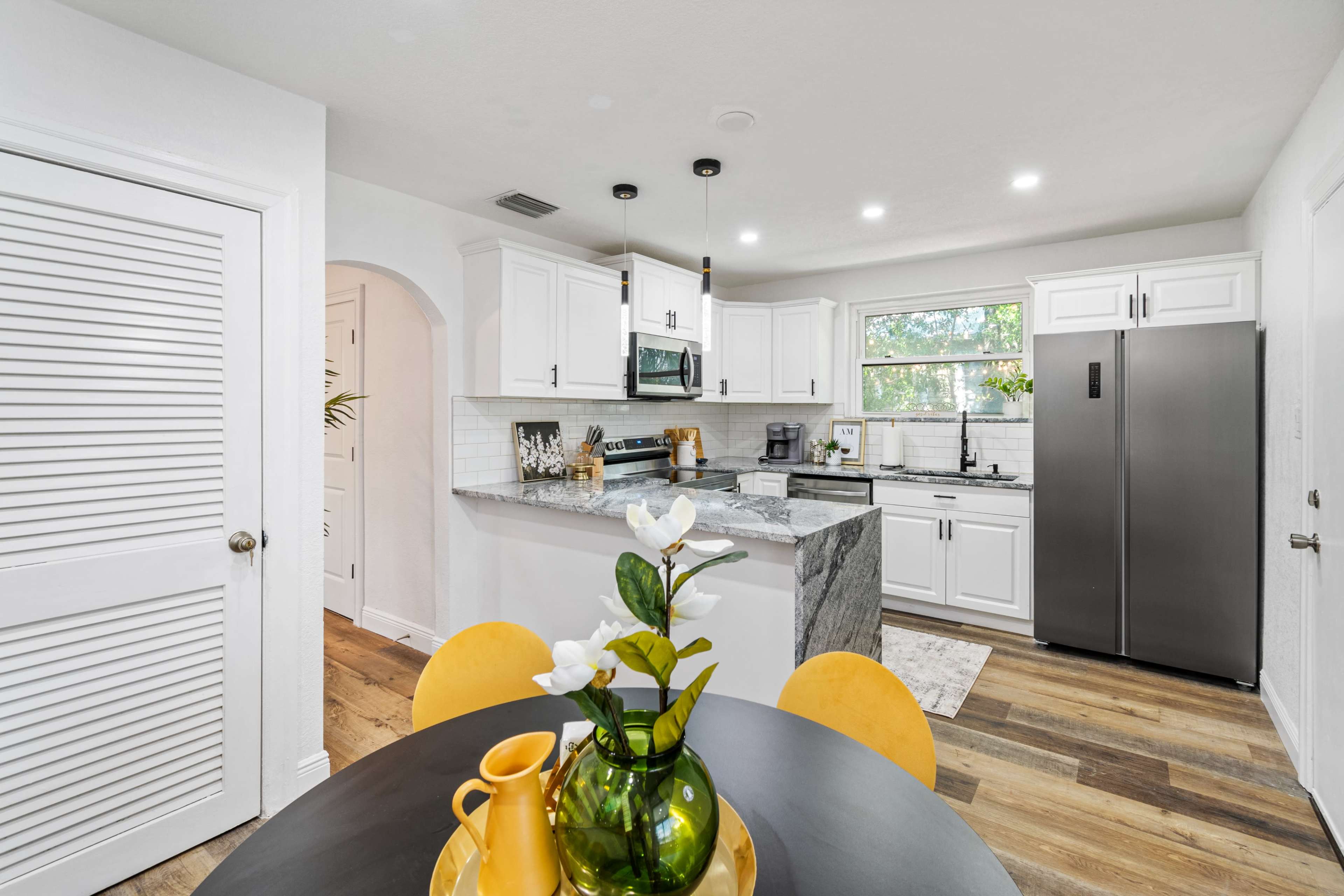 A modern kitchen features white cabinetry, a marble countertop, stainless steel appliances, and a round dining table with yellow chairs.