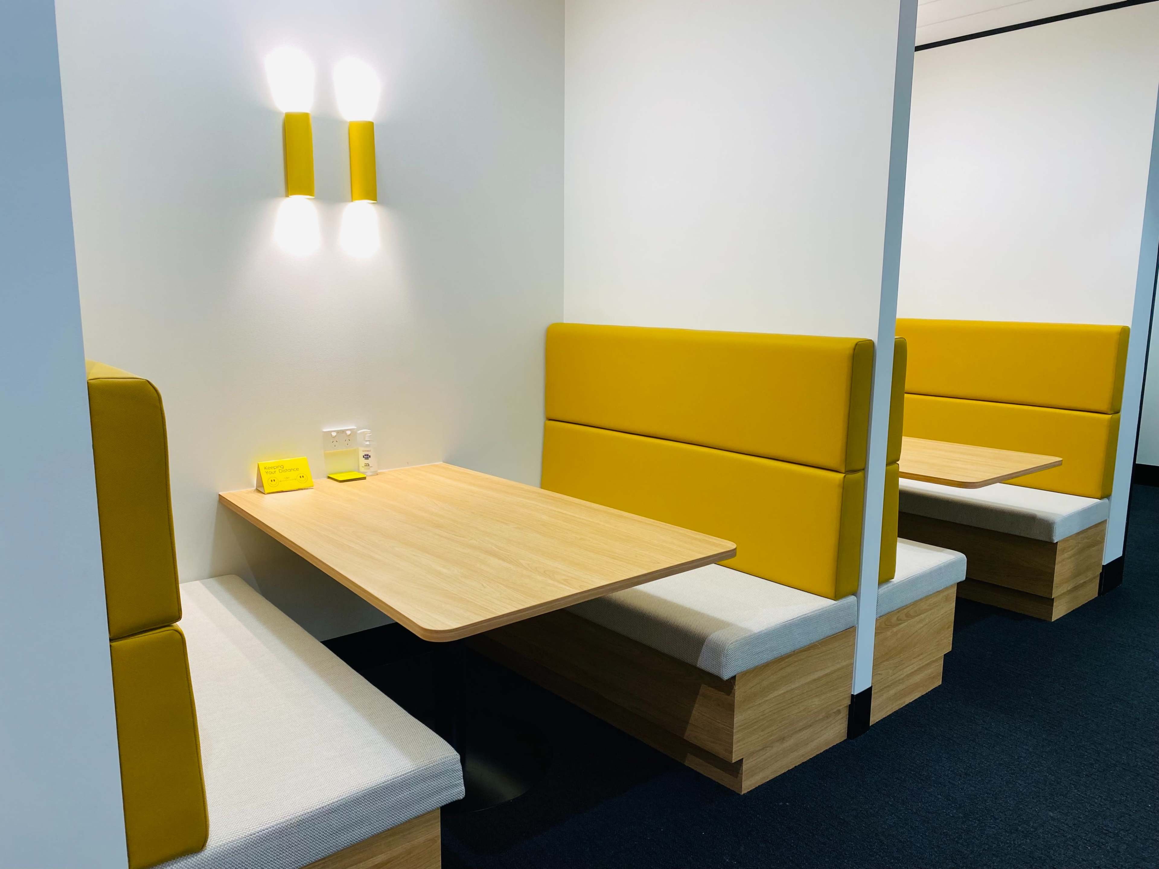 The image shows a modern seating area with yellow-accented booths and a wooden table in a brightly lit space.