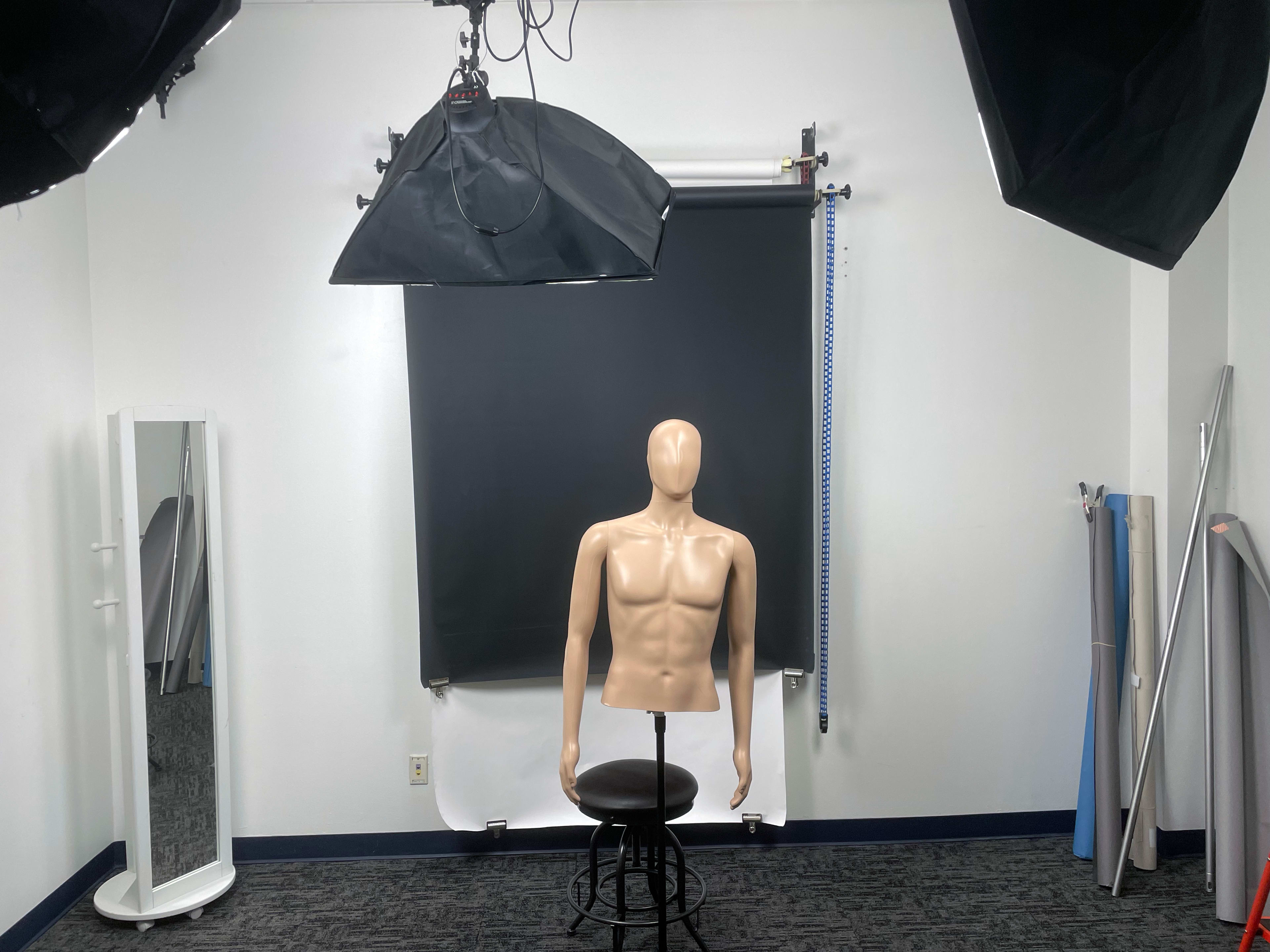 A mannequin with a torso sits on a stool in a photography studio featuring black and white backdrops and lighting equipment.