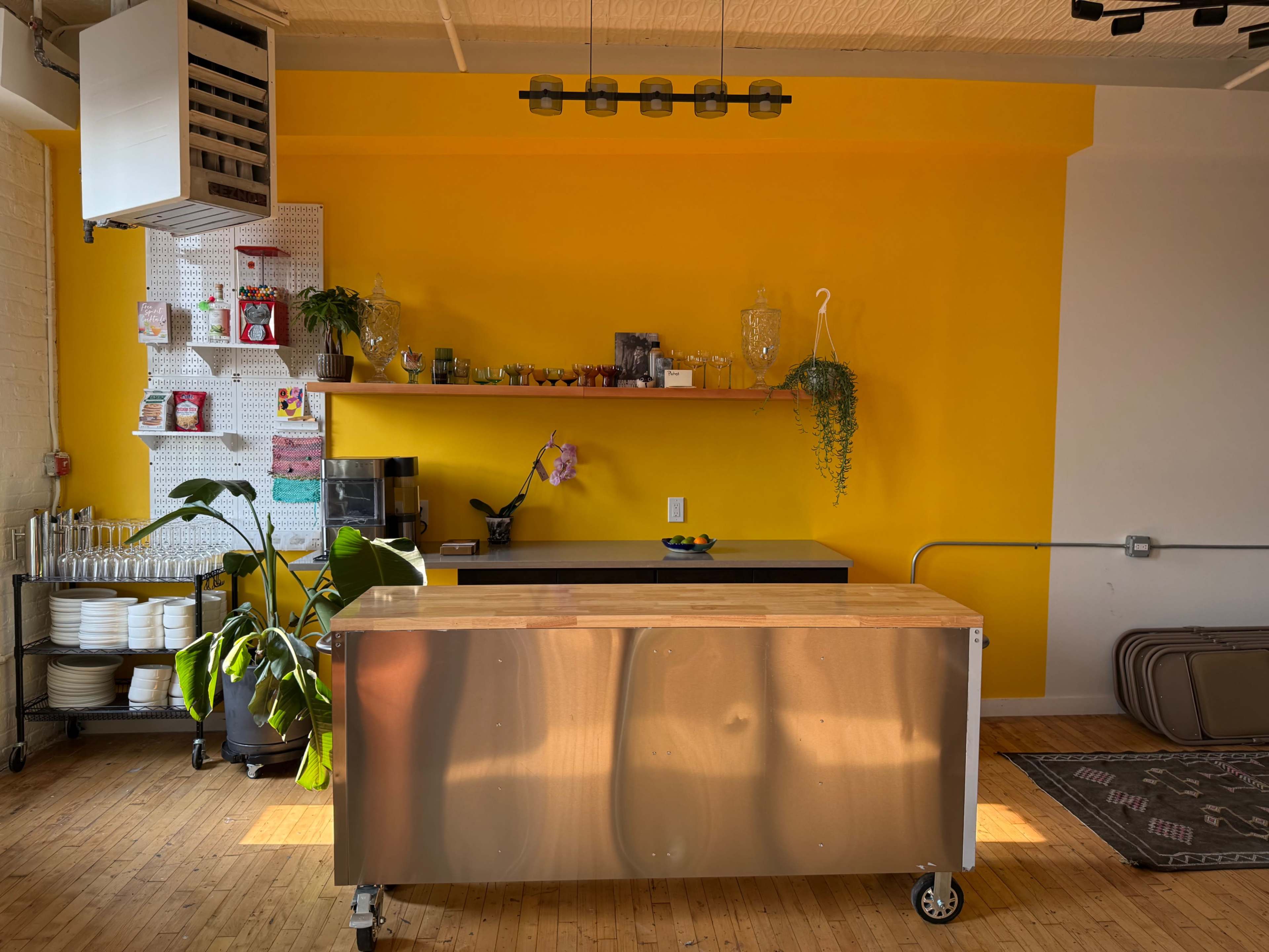 The image shows a bright yellow kitchen with a wooden countertop, a shelf displaying plants and kitchen items, and various dishes arranged on the side.