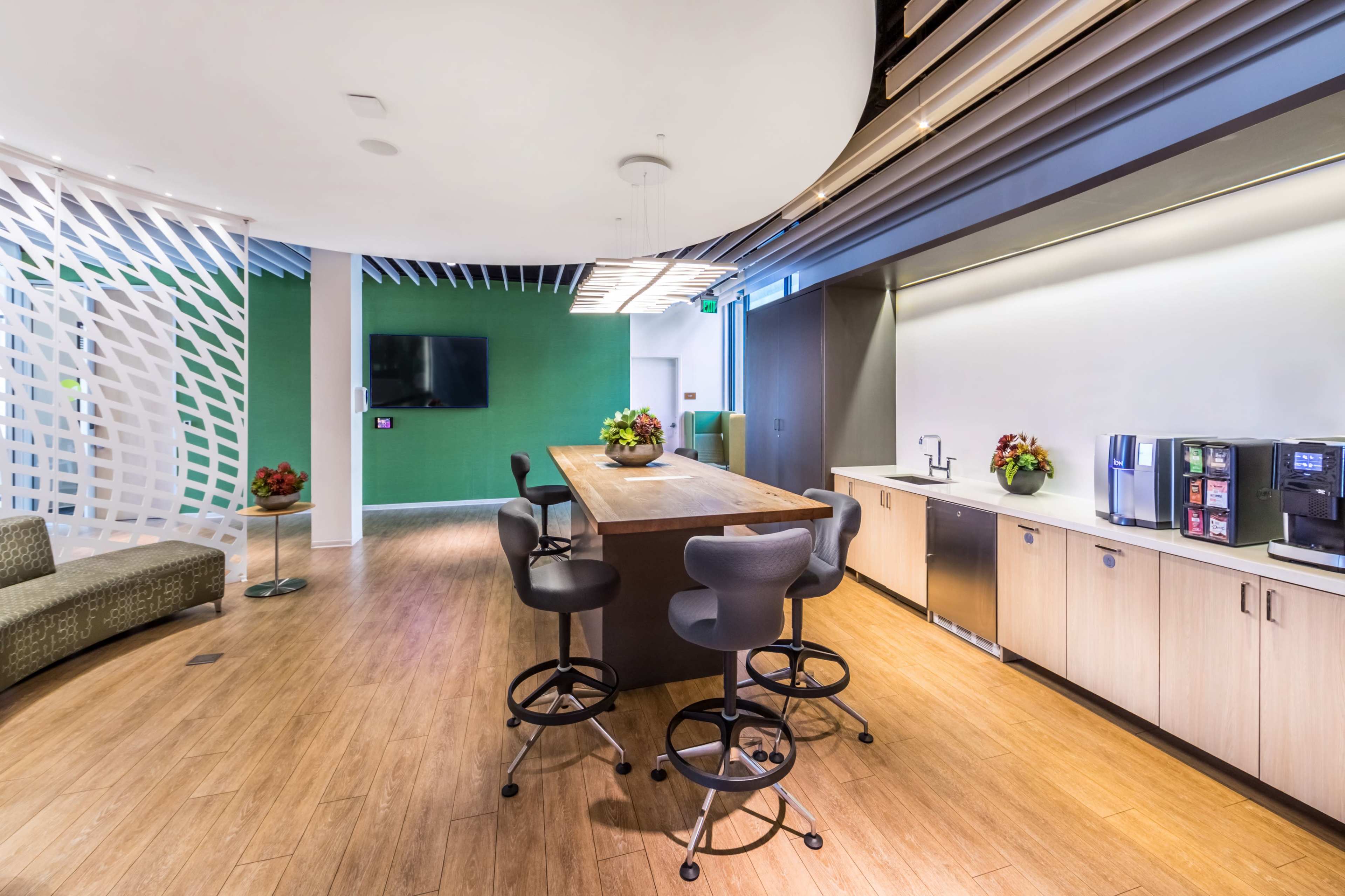 A modern break room with a large communal table, seating, a kitchenette, and a television mounted on the wall.