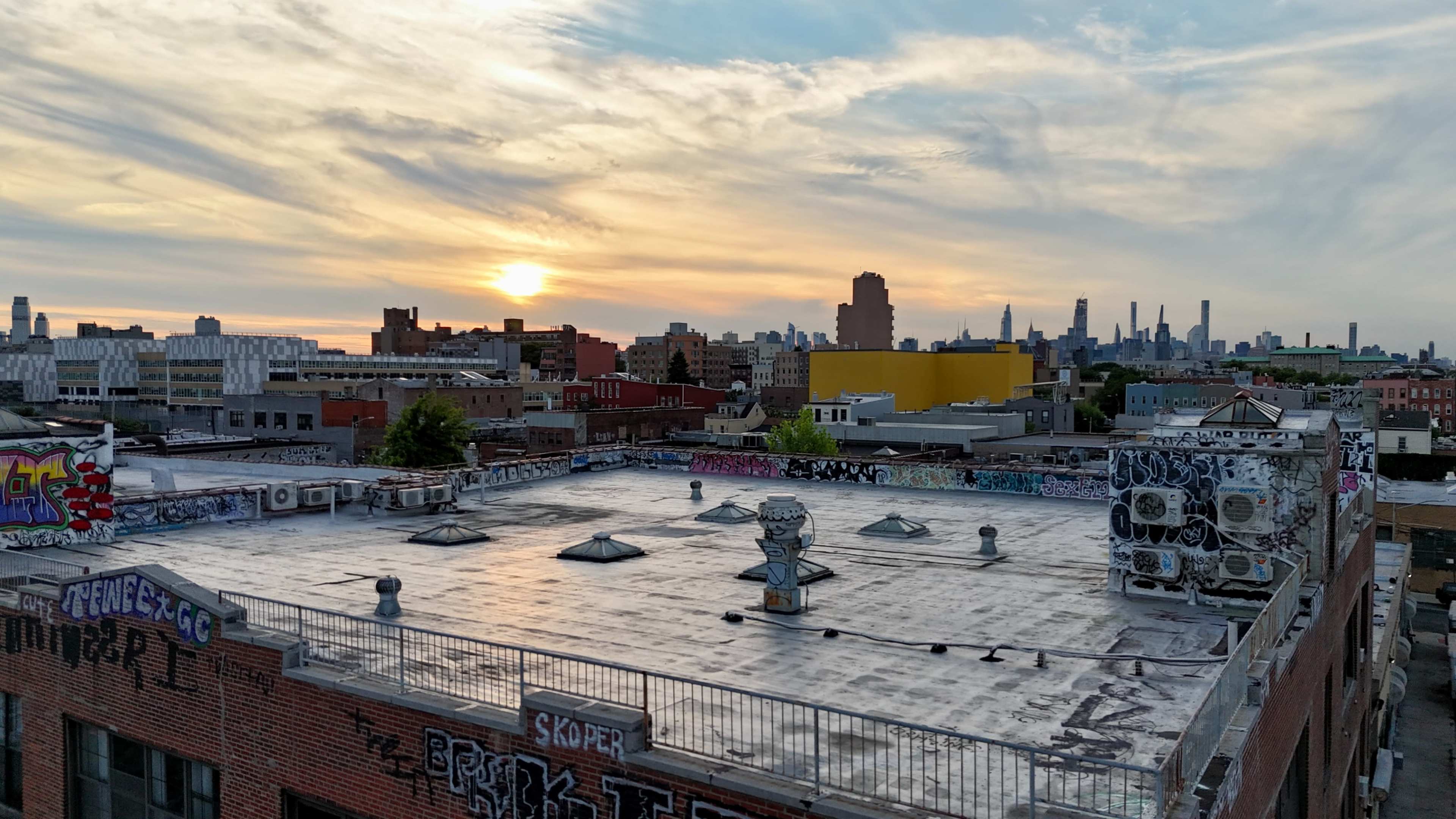 A city rooftop with graffiti-covered walls against a sunset skyline featuring various buildings in the background.