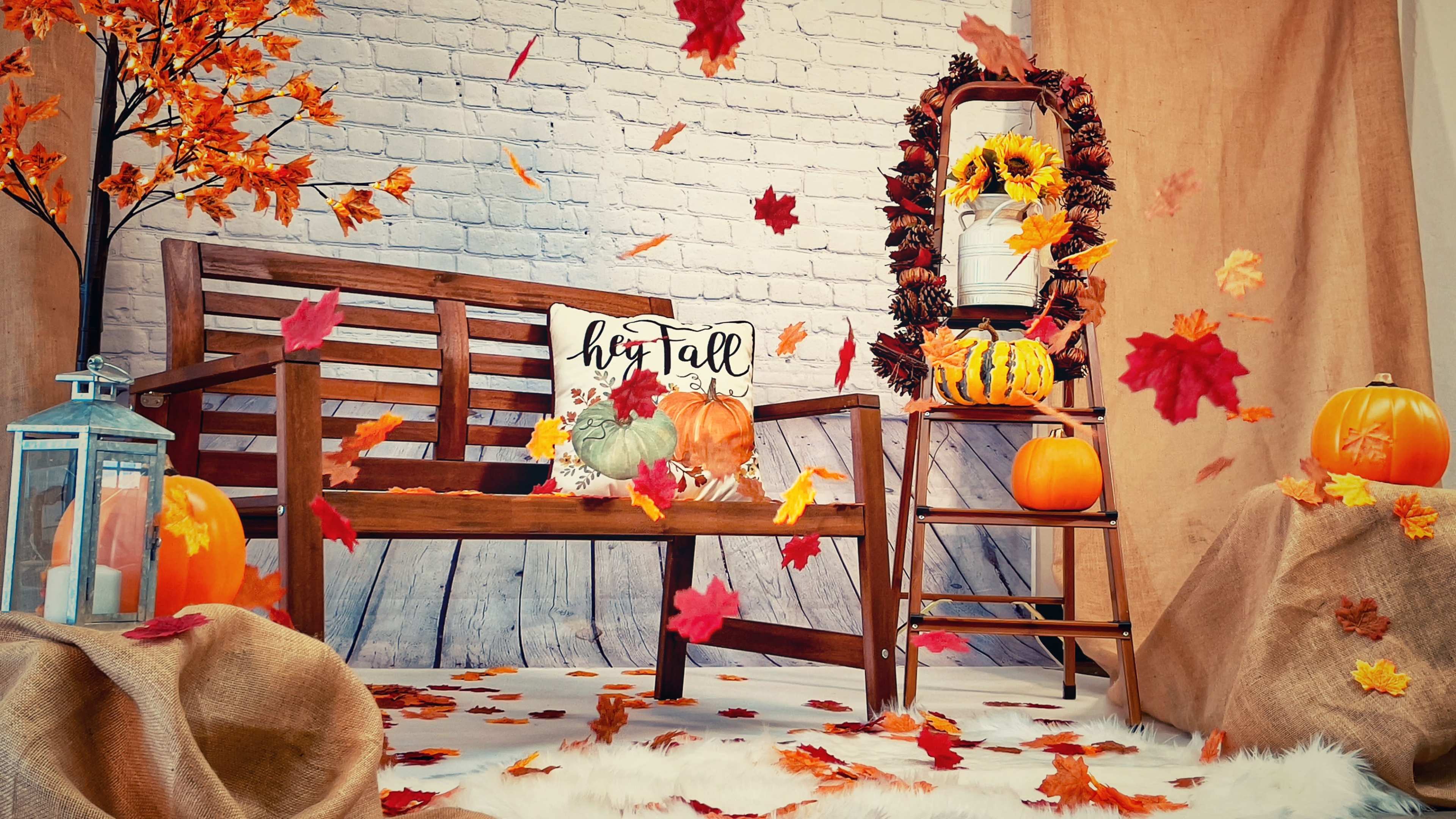 A wooden bench surrounded by autumn-themed decorations, including pumpkins, a sunflower wreath, and artificial falling leaves, creates a seasonal scene.