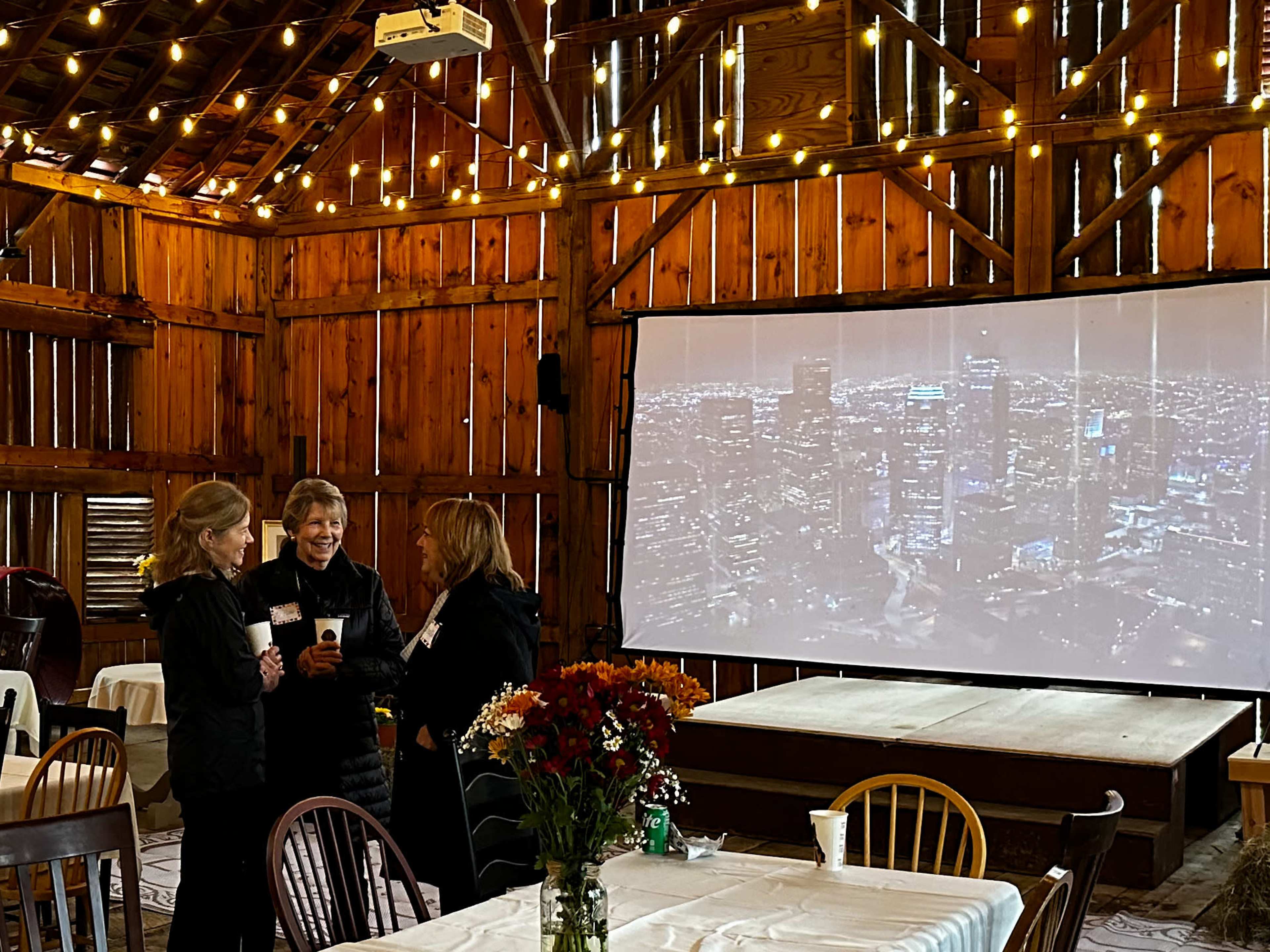 Three women converse in a rustic barn decorated with string lights and a large projection screen displaying a city skyline.