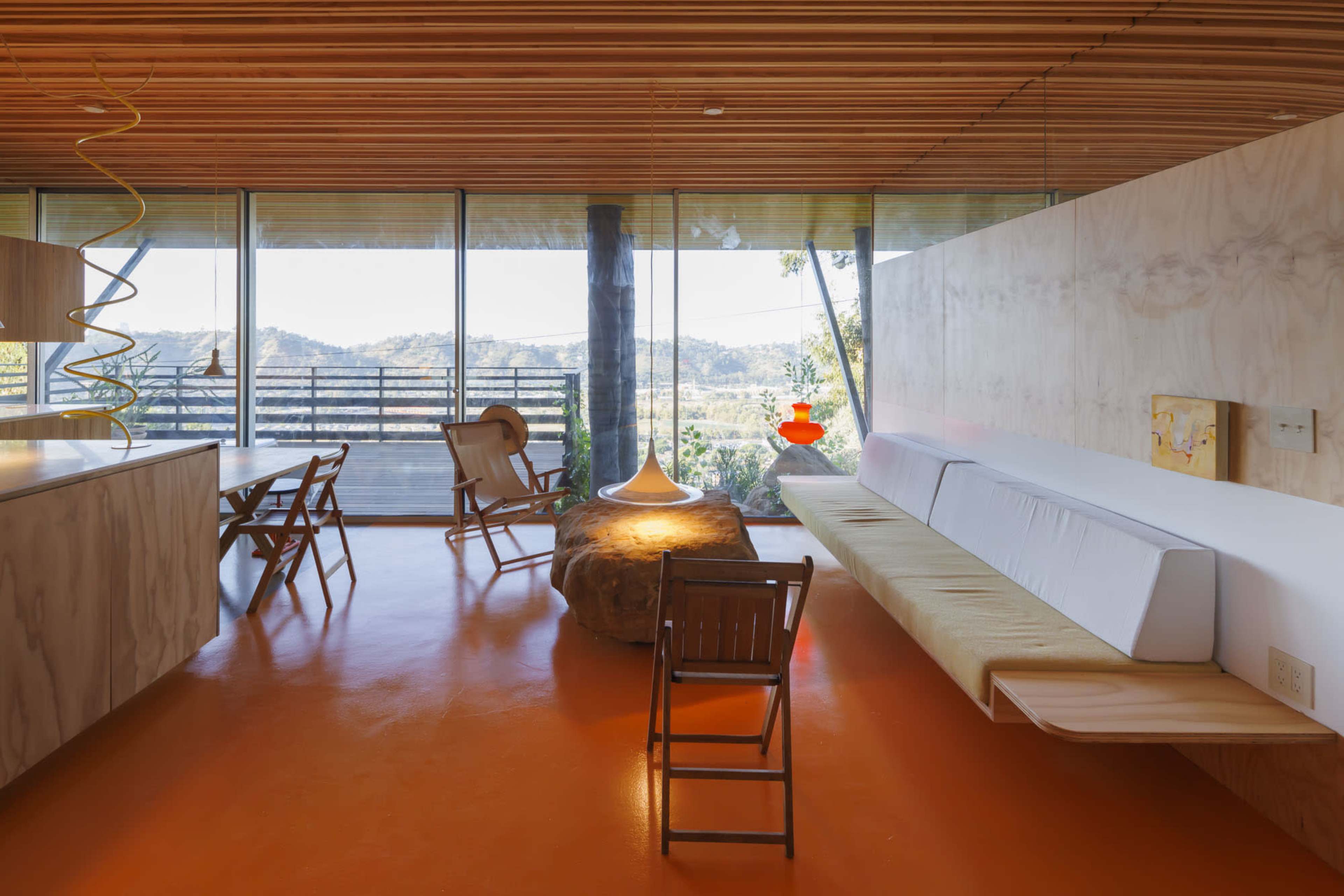 The interior features a modern open space with wooden ceilings, large glass windows showcasing a view of the outdoors, and minimalist furniture arranged around a central table.