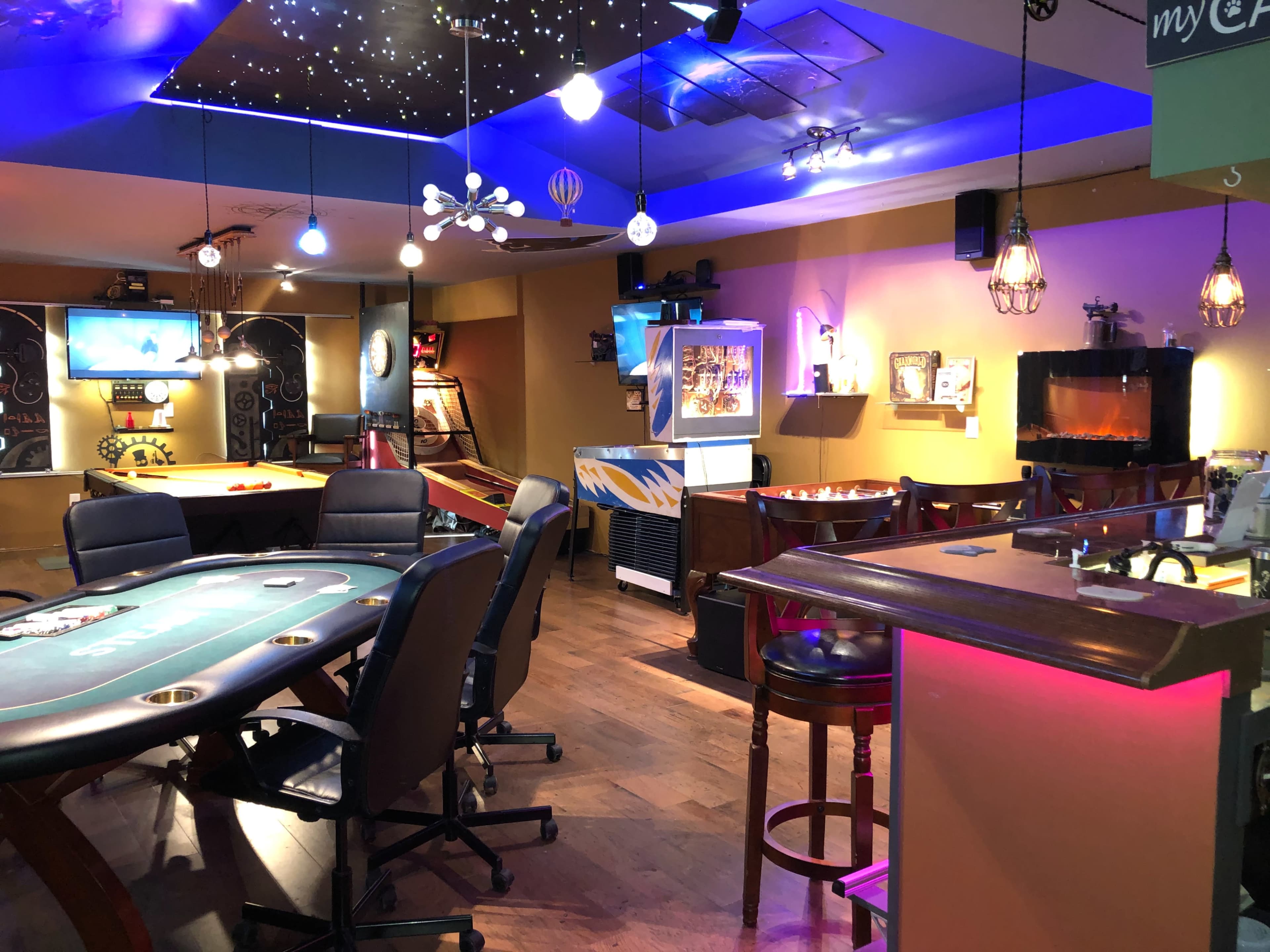 A vibrant gaming and entertainment area featuring a poker table, arcade machine, bar setup, and various seating arrangements under colorful lighting.