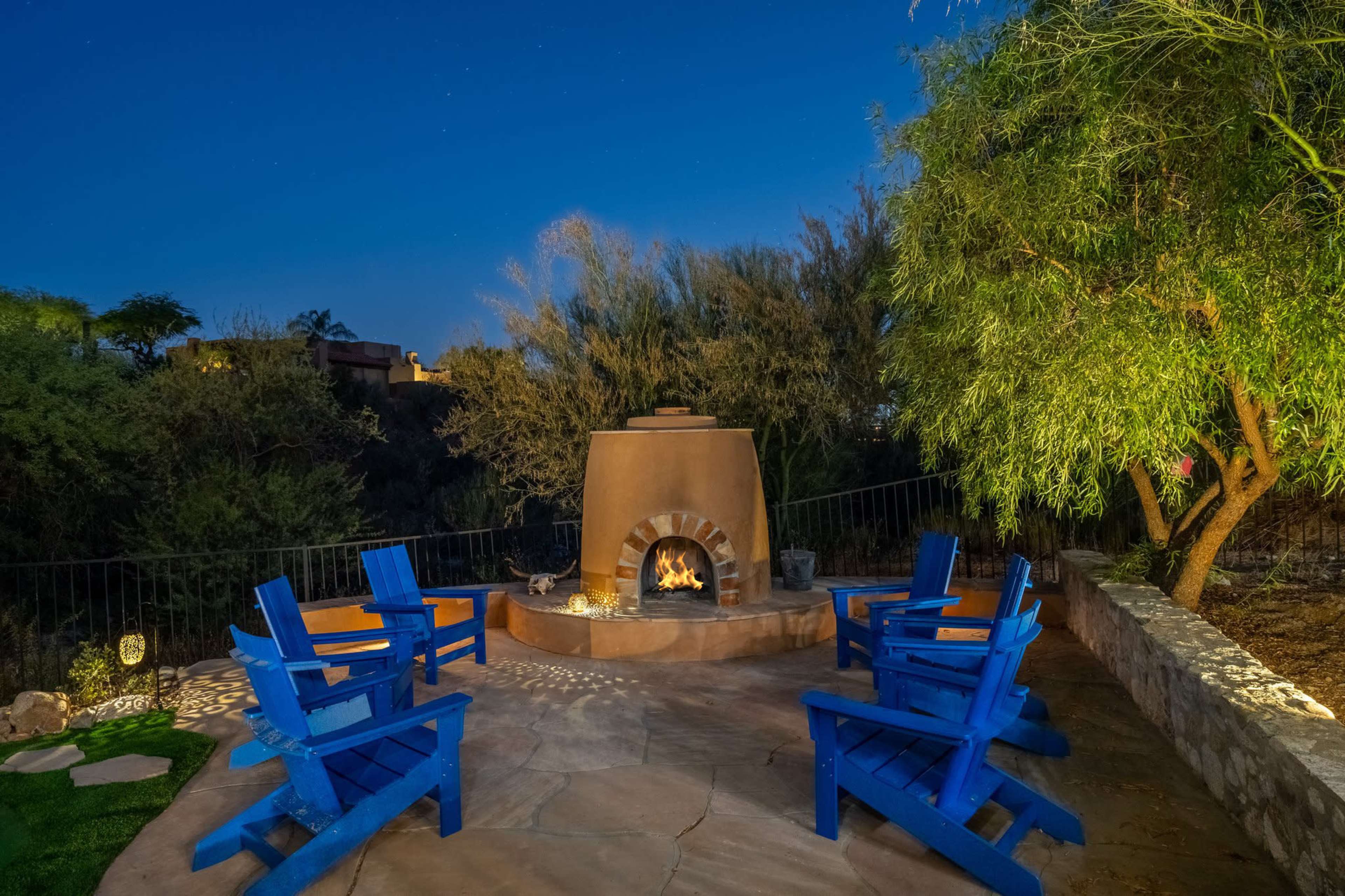 A circular stone patio features a brown chiminea surrounded by four blue Adirondack chairs under a night sky.