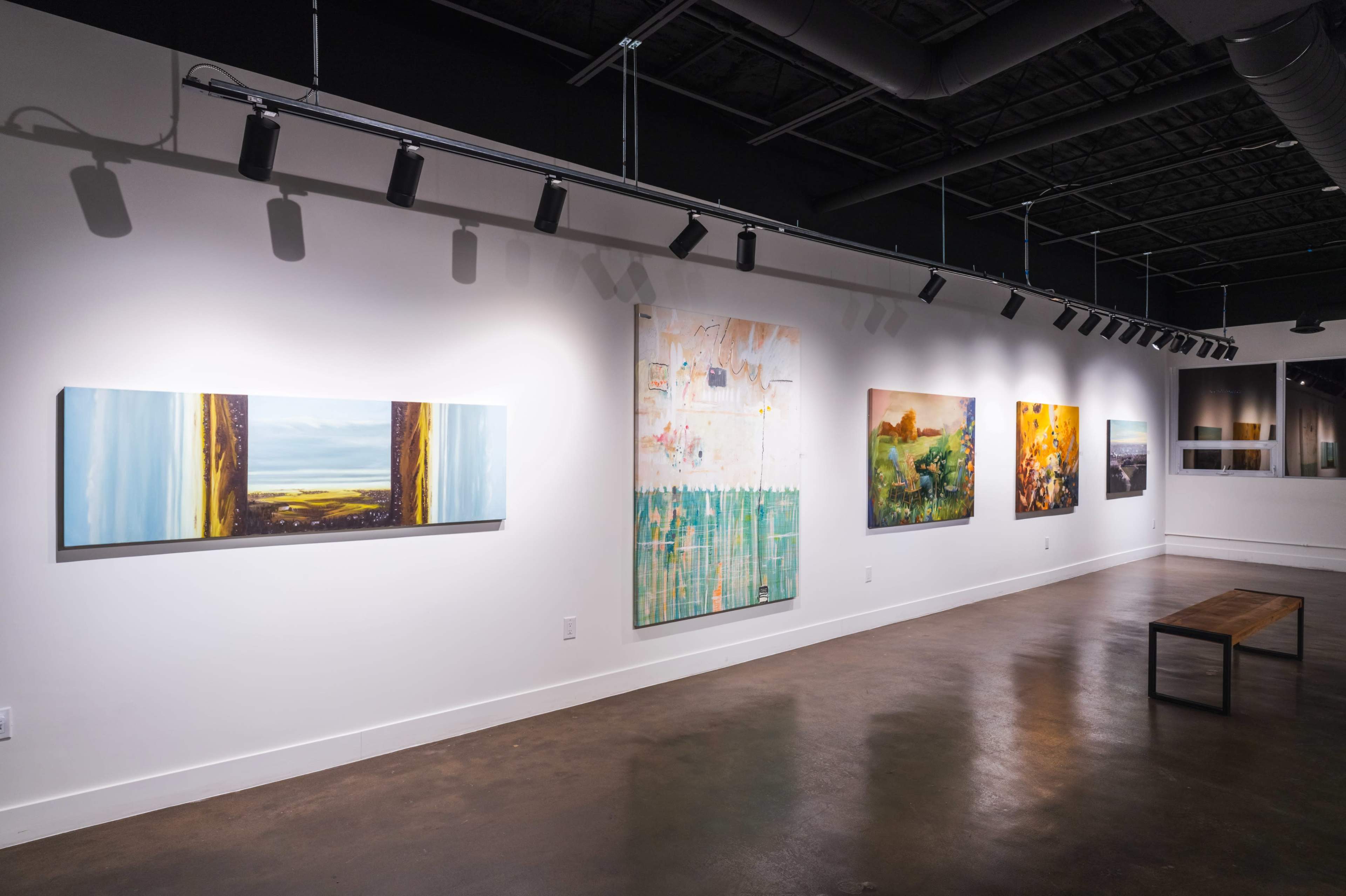 The image shows an art gallery with a series of colorful paintings displayed along a white wall, accompanied by a wooden bench in the center of the room.