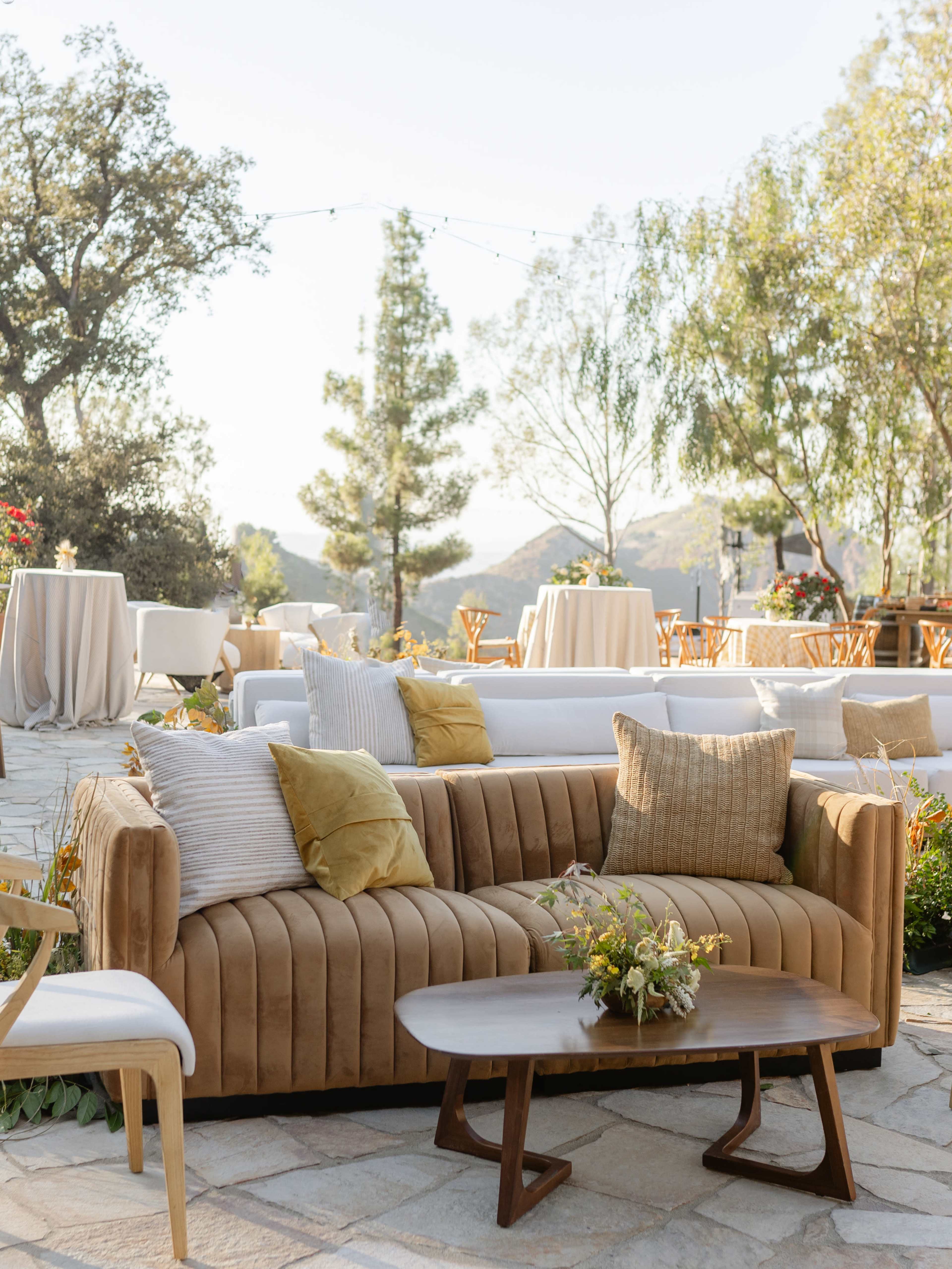 A stylish outdoor seating area features a brown couch with decorative pillows, a small wooden table, and various tables set for an event amidst greenery and mountains in the background.