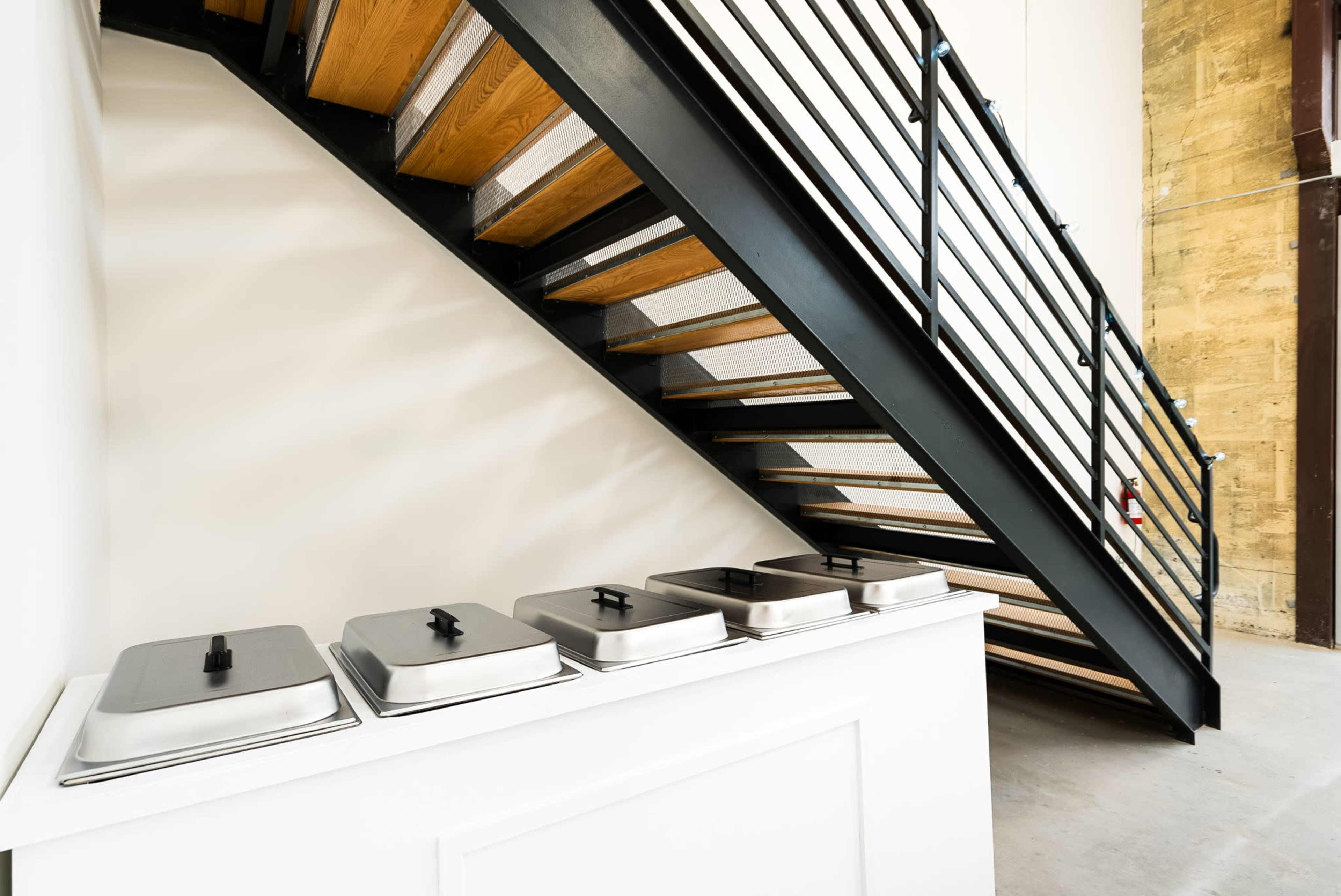 A row of metallic food warmers is positioned under a staircase with wooden steps and a black railing.