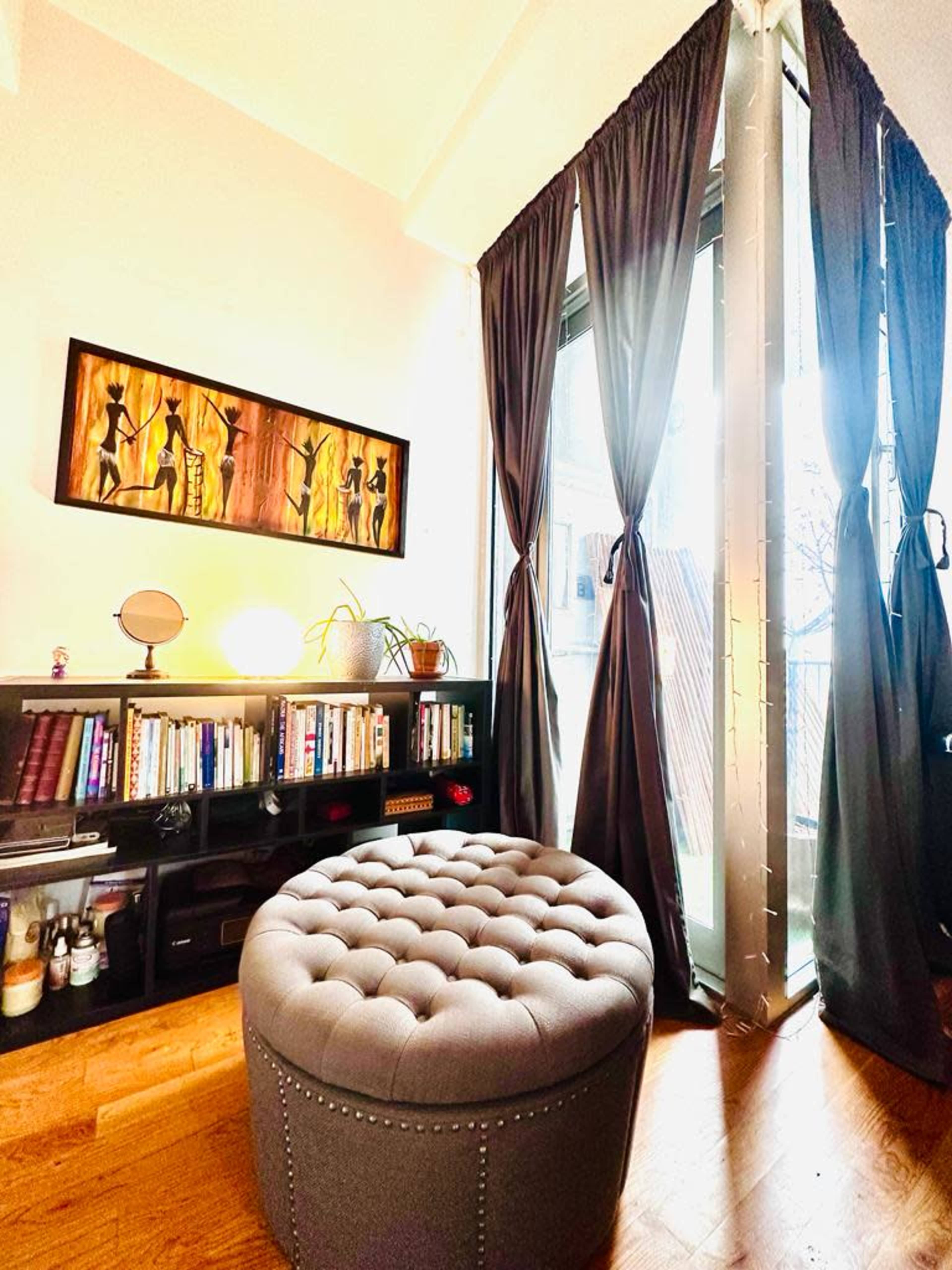 The image shows a cozy room featuring a tufted ottoman, a bookshelf filled with books, and floor-to-ceiling curtains beside a large window.