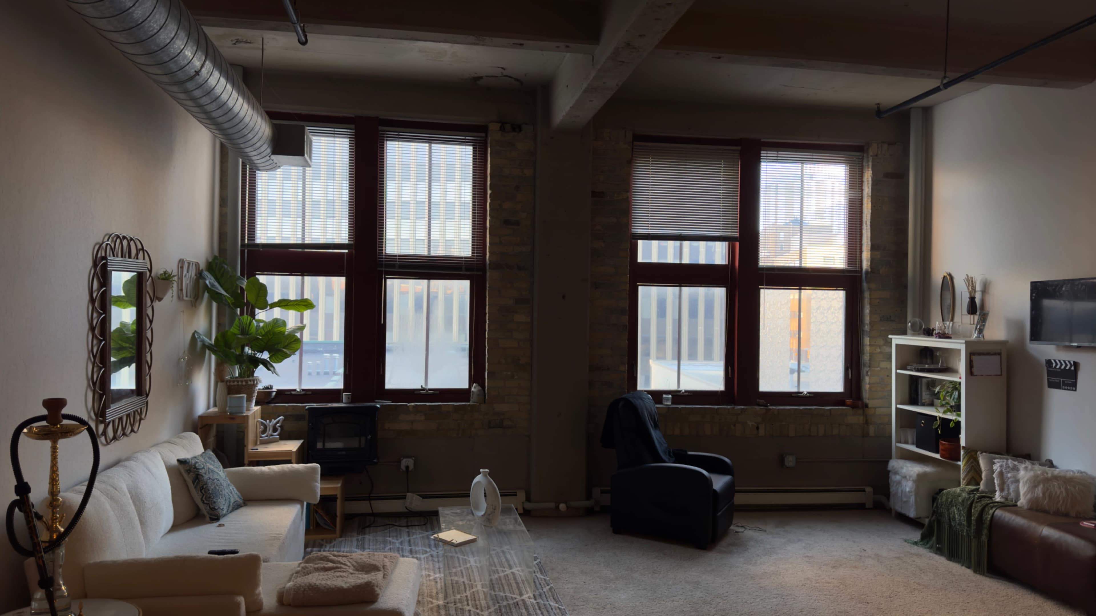 A bright room features large windows, a white couch, a black chair, and a few decorative elements against a brick wall.