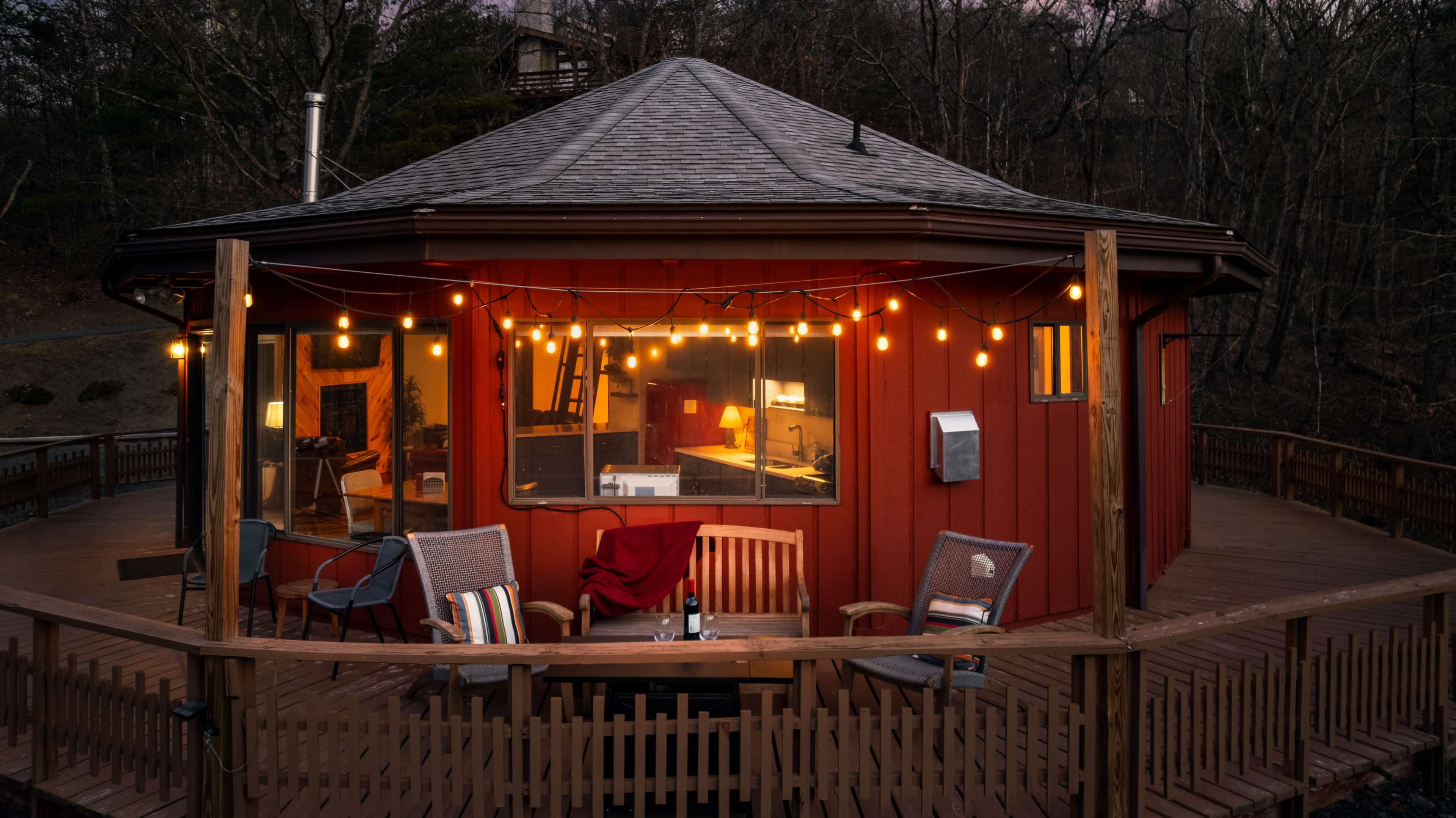 A round, red cabin with a wooden deck is illuminated by string lights, surrounded by trees in a twilight setting.