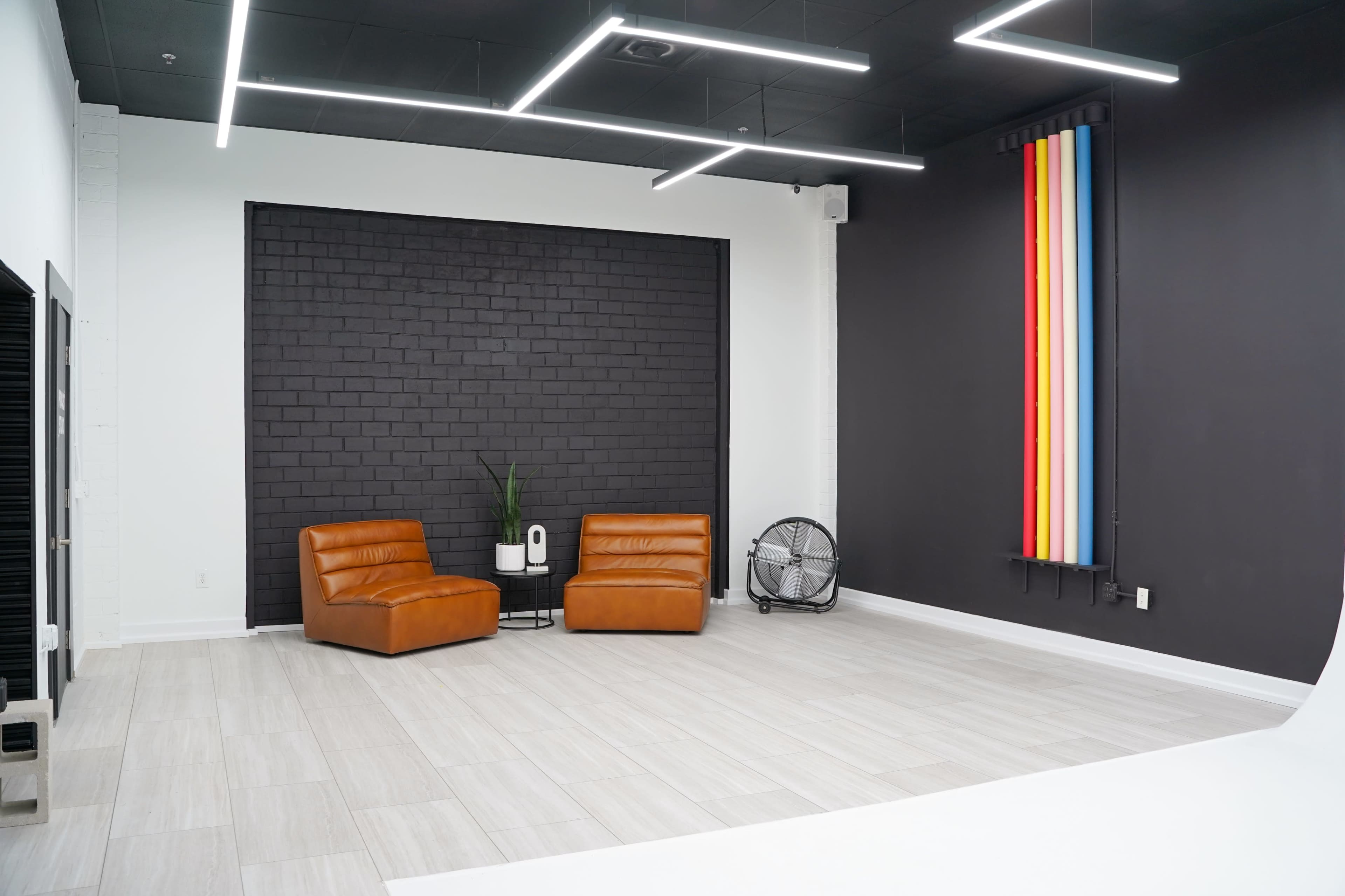 A modern studio space with two brown chairs, a small table, a potted plant, and colorful vertical strips against a black wall.
