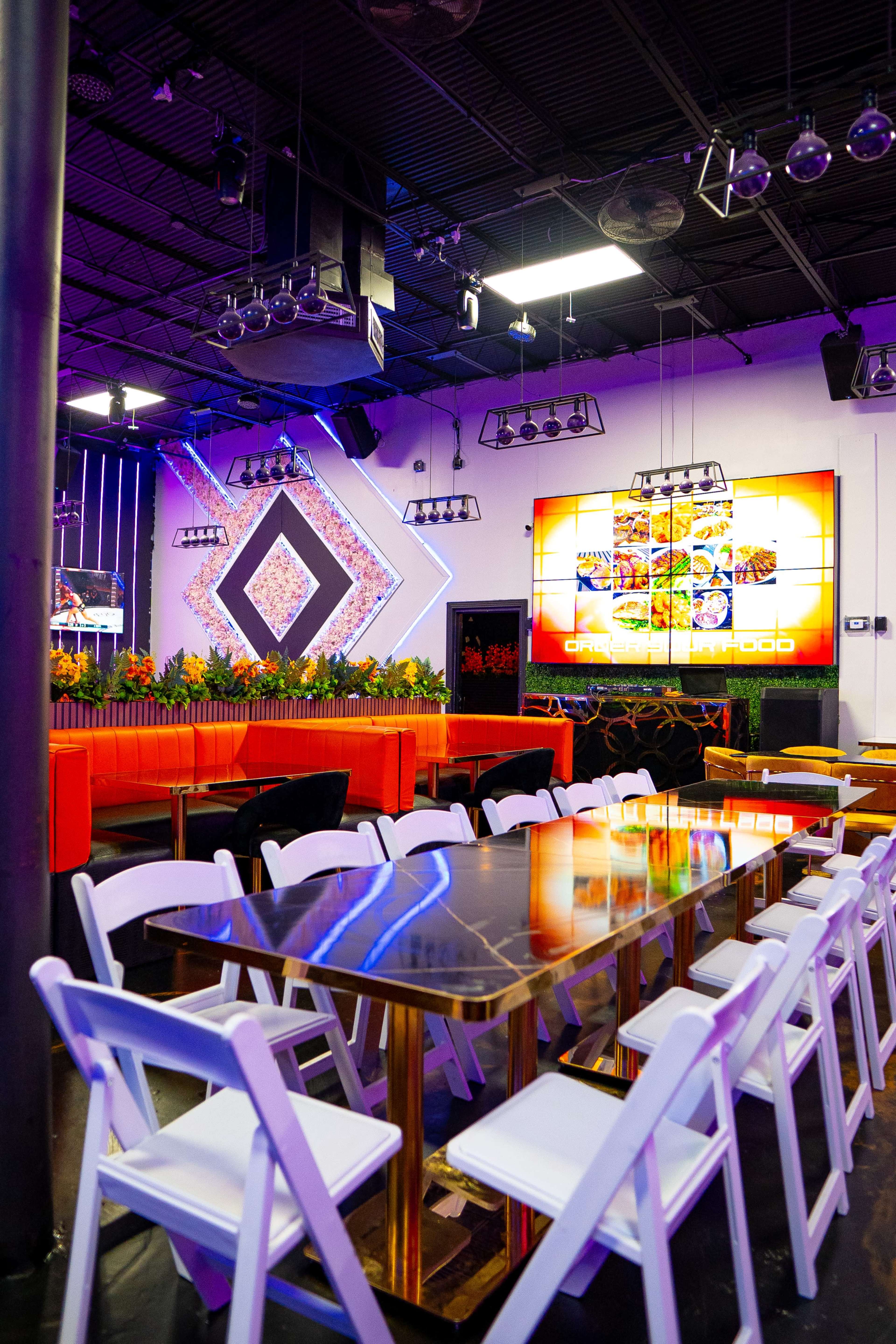 The image shows a modern interior of a restaurant or event space with colorful lighting, a large screen displaying food images, and seating arrangements featuring white chairs and a black table.