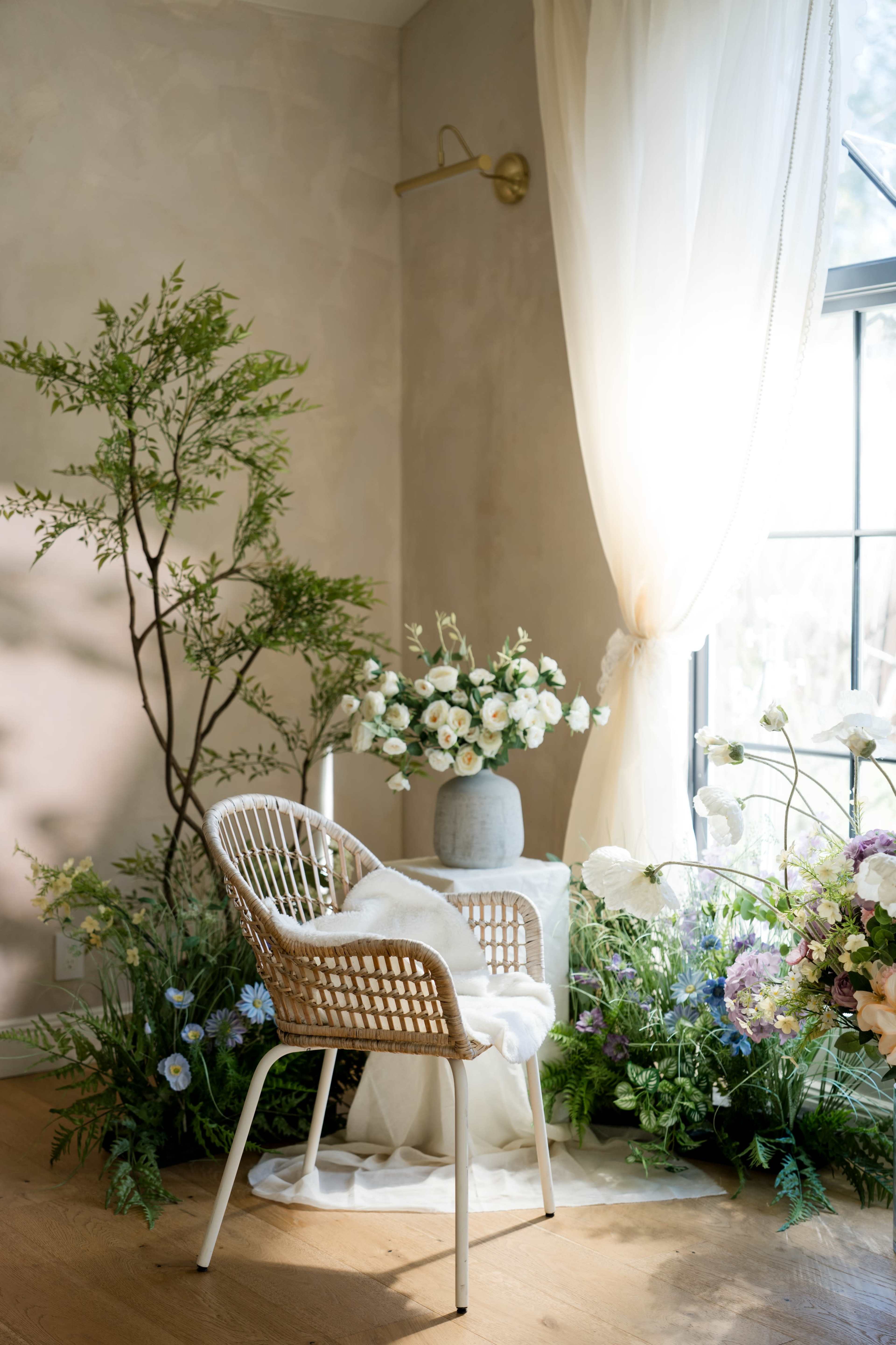 A chair surrounded by a variety of flowers and greenery sits near a window with soft, flowing curtains.