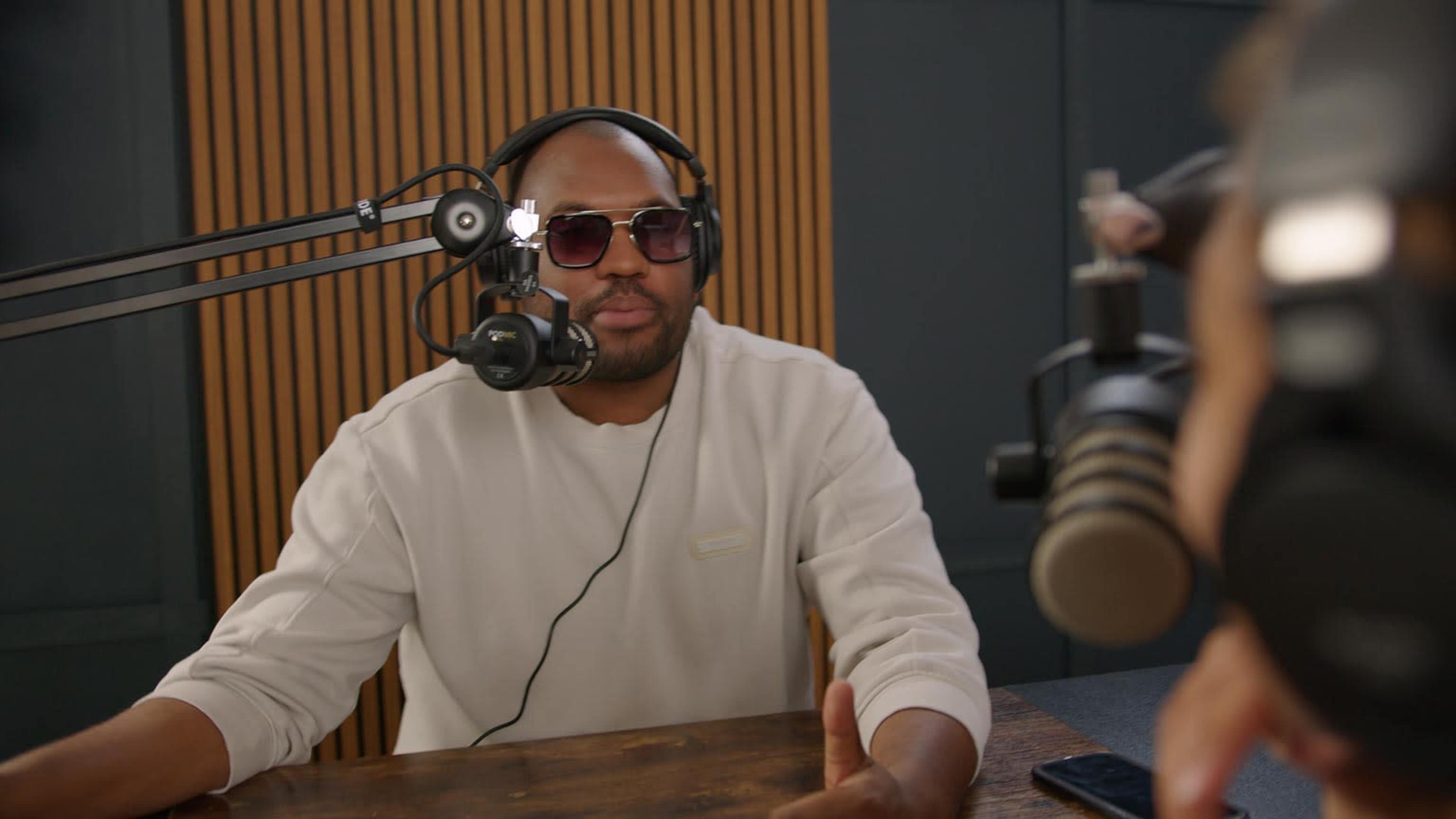 A man wearing sunglasses and headphones speaks into a microphone in a recording studio.